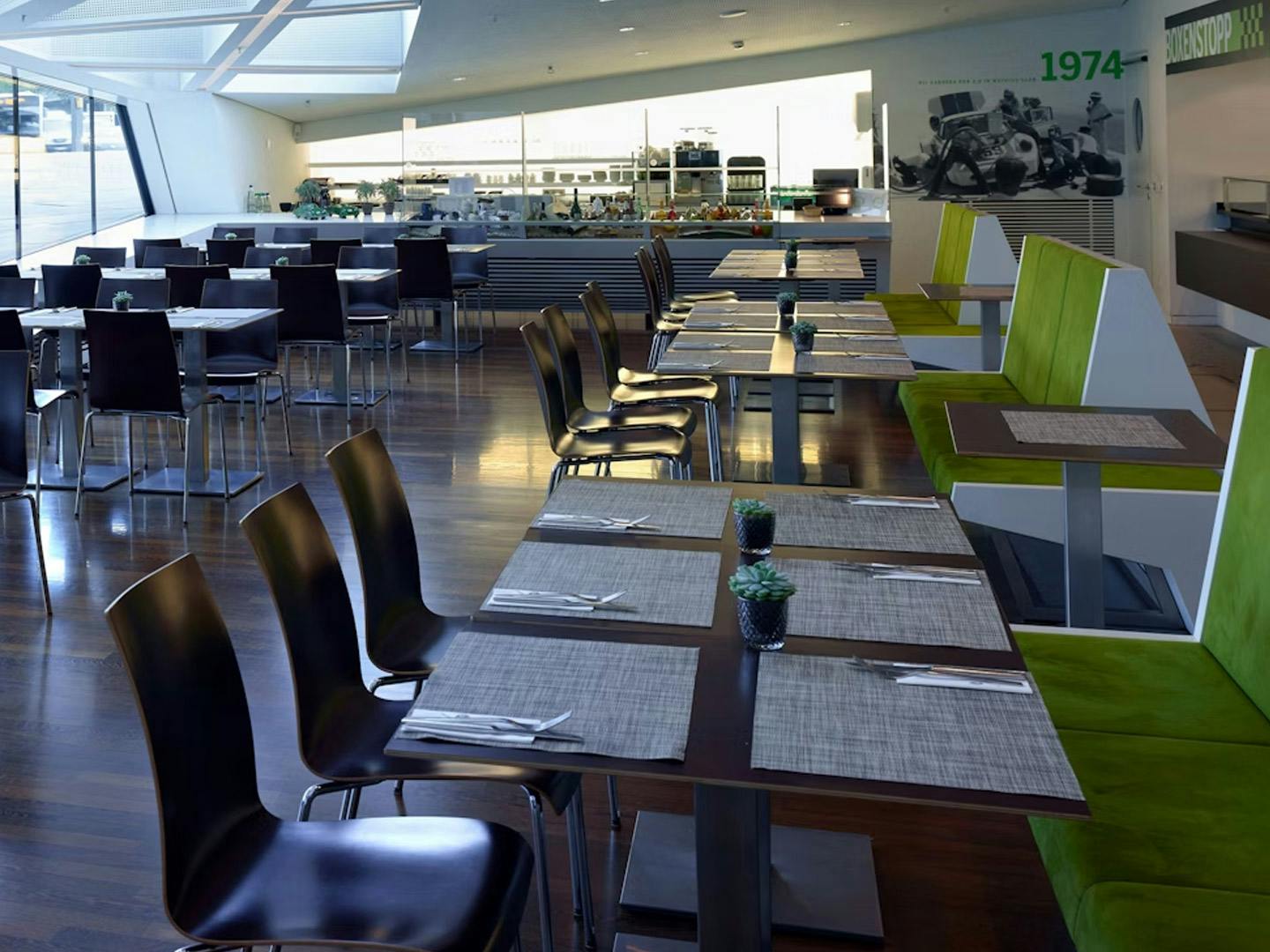 Das moderne helle Porsche Bistro Boxenstopp mit mehreren Tischreihen und Stühlen. Rechts im Bild ist eine grün gepolsterte Sitzreihe.