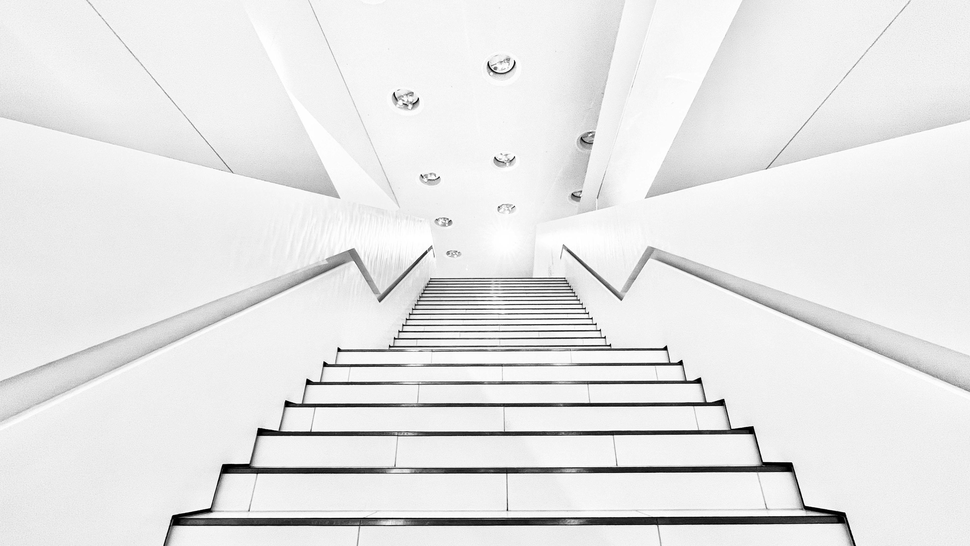 Symmetrische Aufnahme einer hellen Museumstreppe mit klaren Linien und runden Deckenleuchten, fotografiert von unten nach oben.