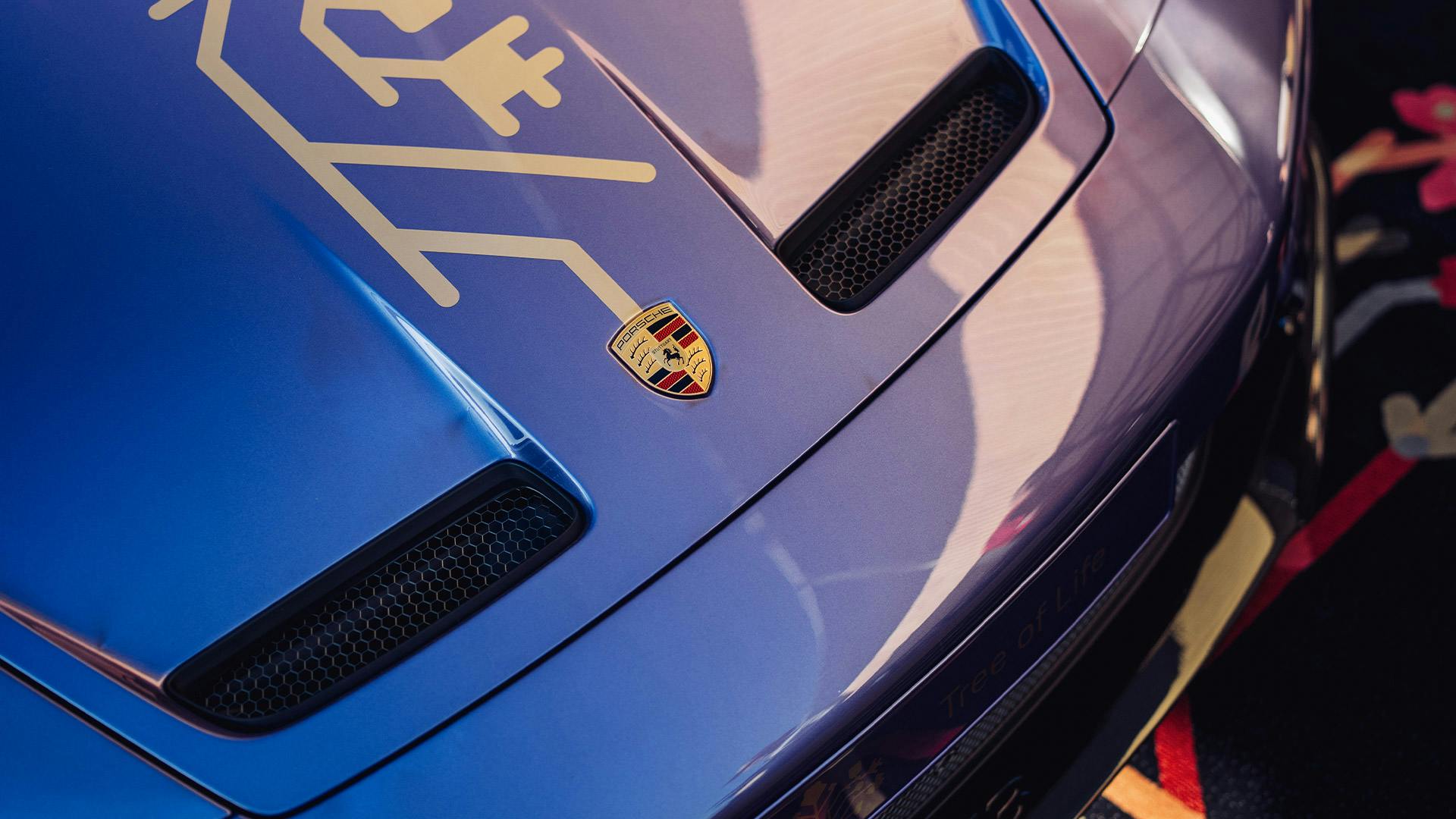 Close-up of a Porsche Tree of Life Sonderwunsch bonnet in metallic blue, featuring a gold crest and decorative gold graphic above the air intakes.