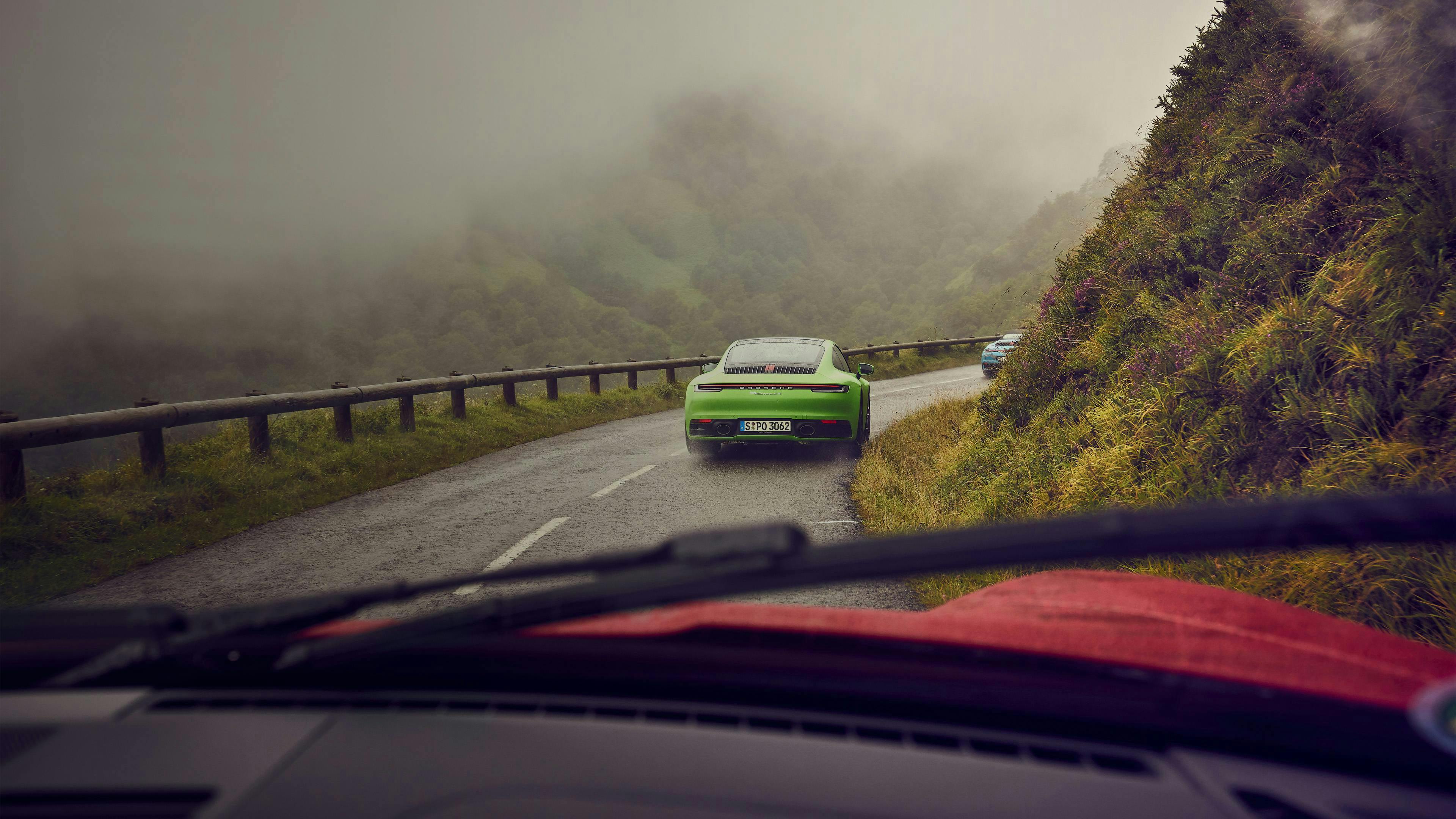 Green Porsche 911 driving through a foggy mountain road, viewed from inside another car, with mist and greenery around.
