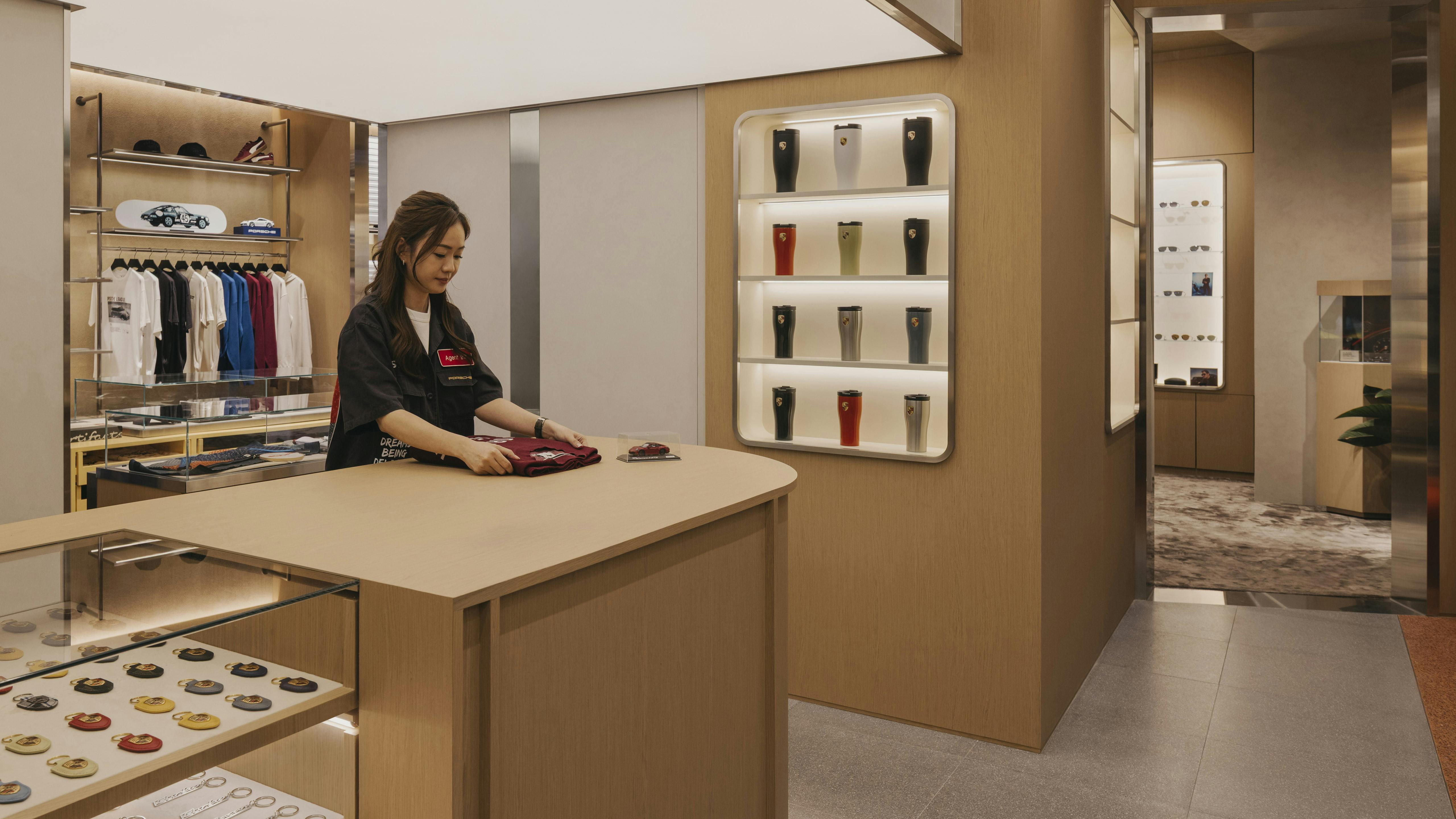 A Porsche at Jewel staff folds a T-shirt in a shot with tumblers and keyrings displayed