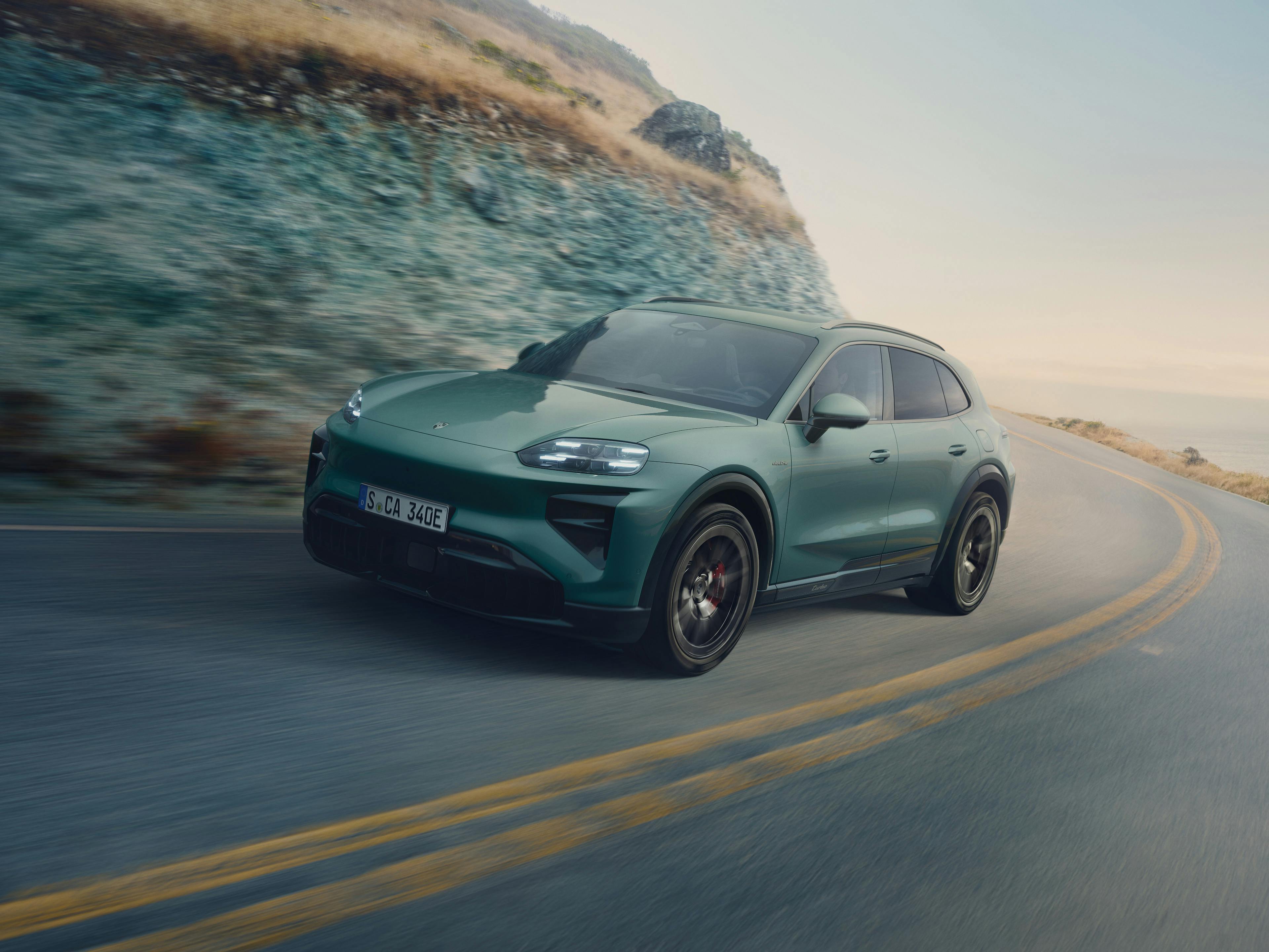 A Mystic Green Metallic Porsche Cayenne Turbo Electric SUV driving on a winding coastal road with rocky cliffs on one side and the sea on the other.