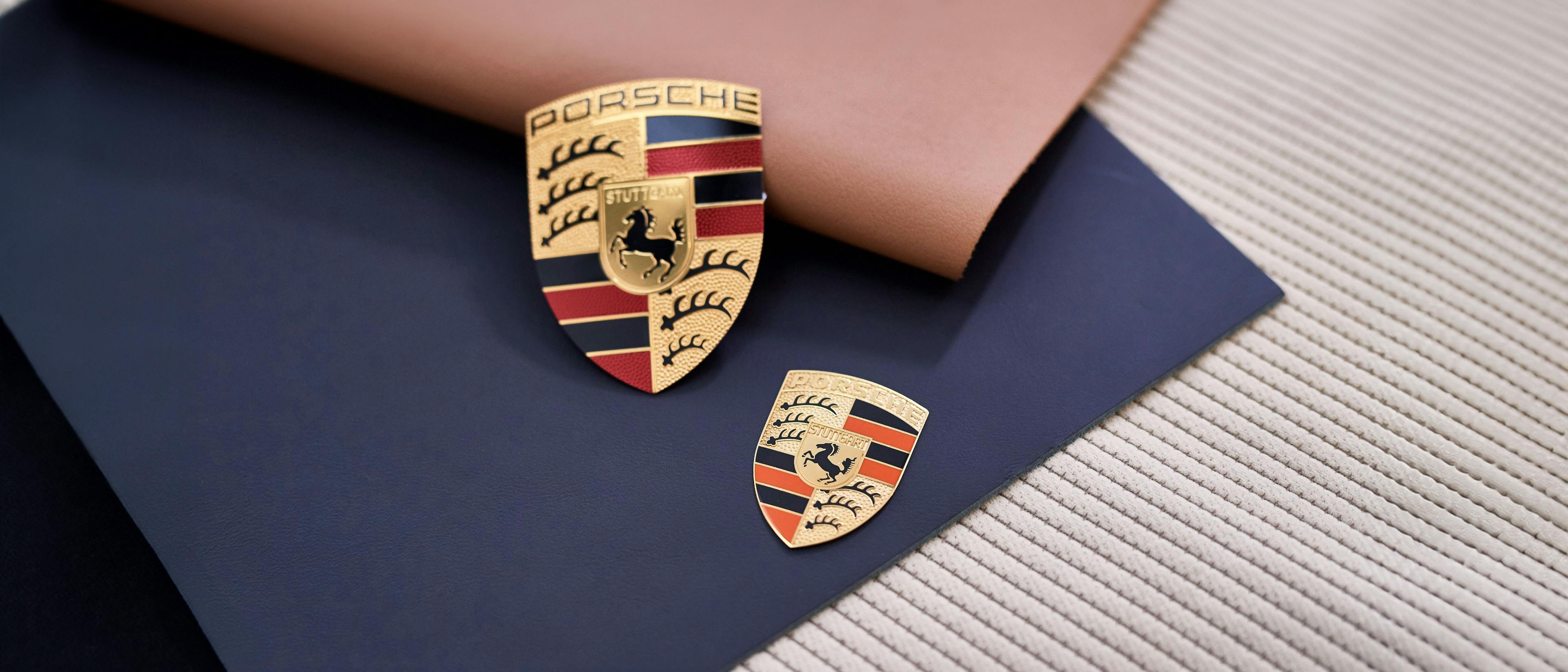 2 Porsche emblemen in verschillende maten liggen op stoffen en lederen patronen.