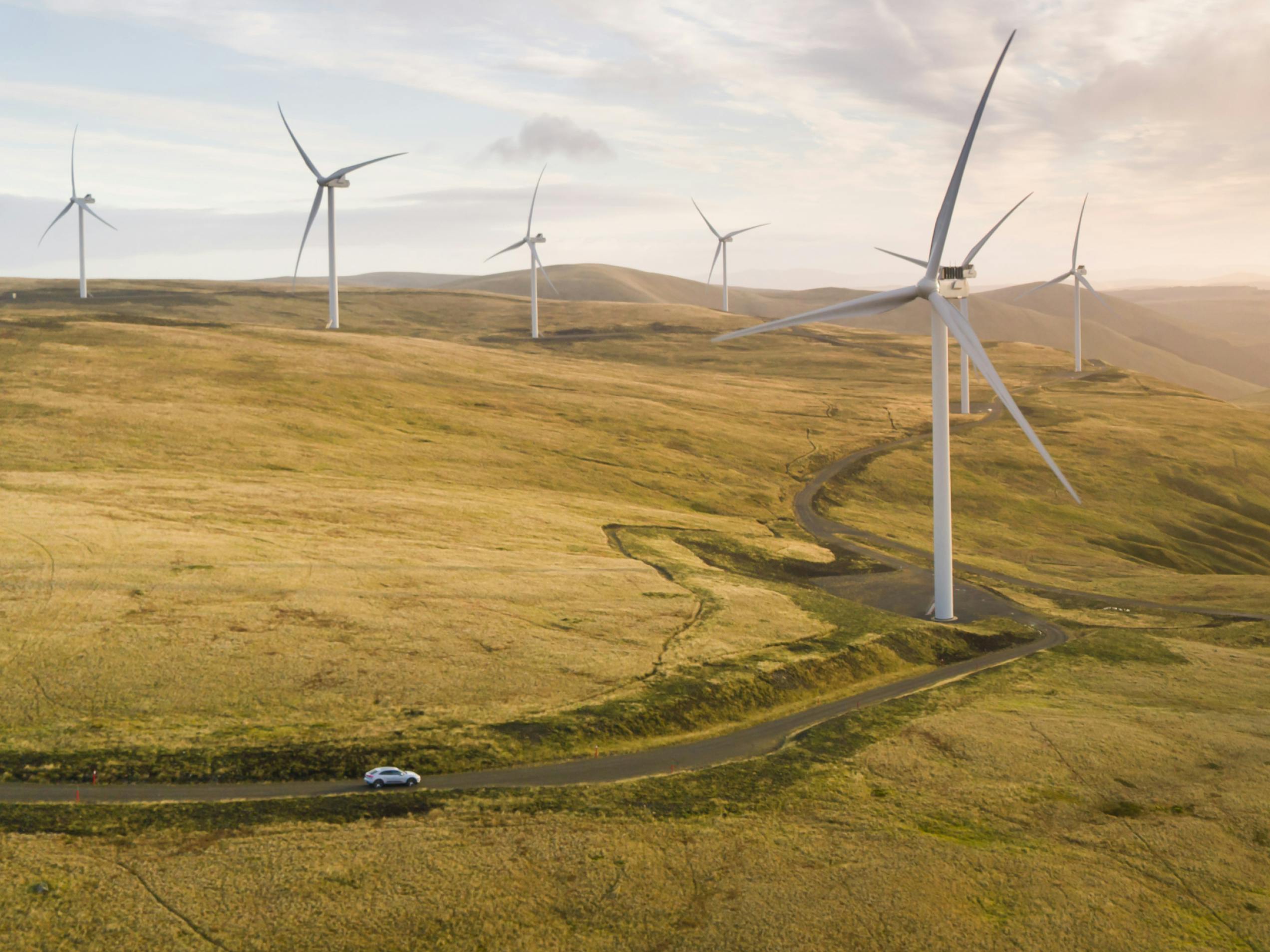 View from above at a distance of a wide, Yellow-Green, hilly field with seven wind turbines. A Porsche is driving past.