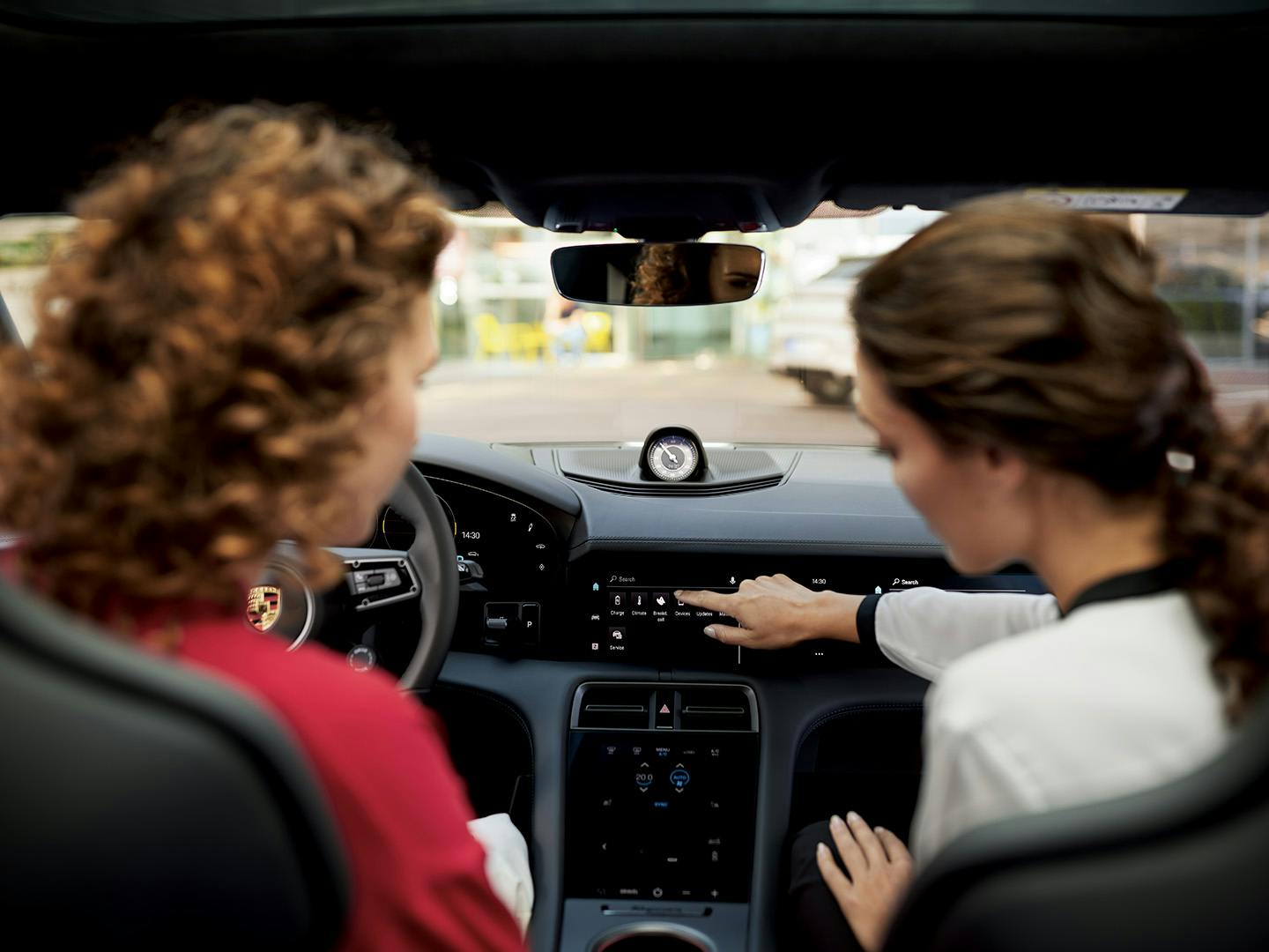 Dos mujeres aparecen en el interior de un Porsche. La mujer del asiento del copiloto señala con el dedo la consola central del vehículo.