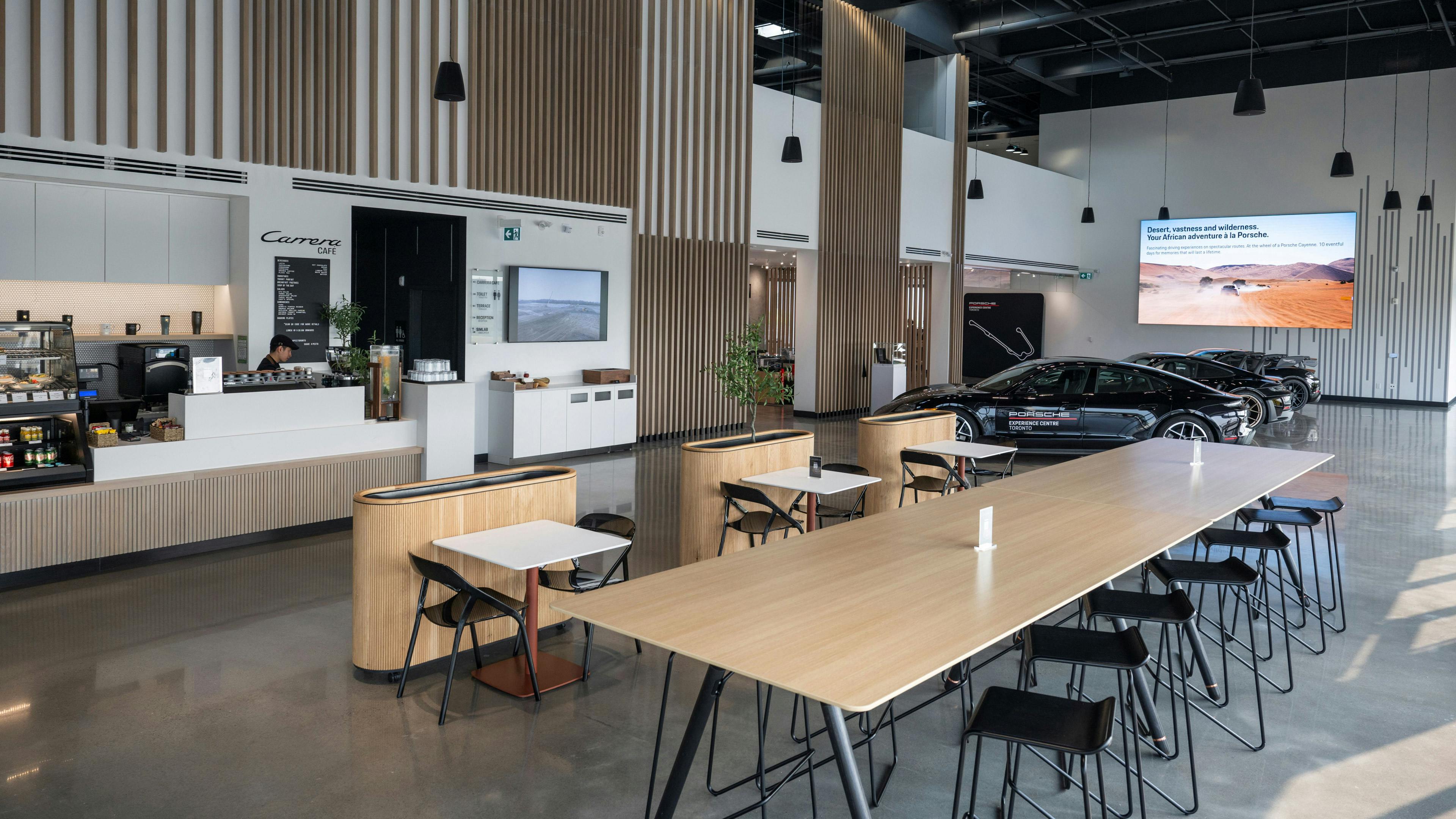 Interior view of a modern lobby with high ceilings, a café area, Porsche vehicles on display, and a large screen in the background.