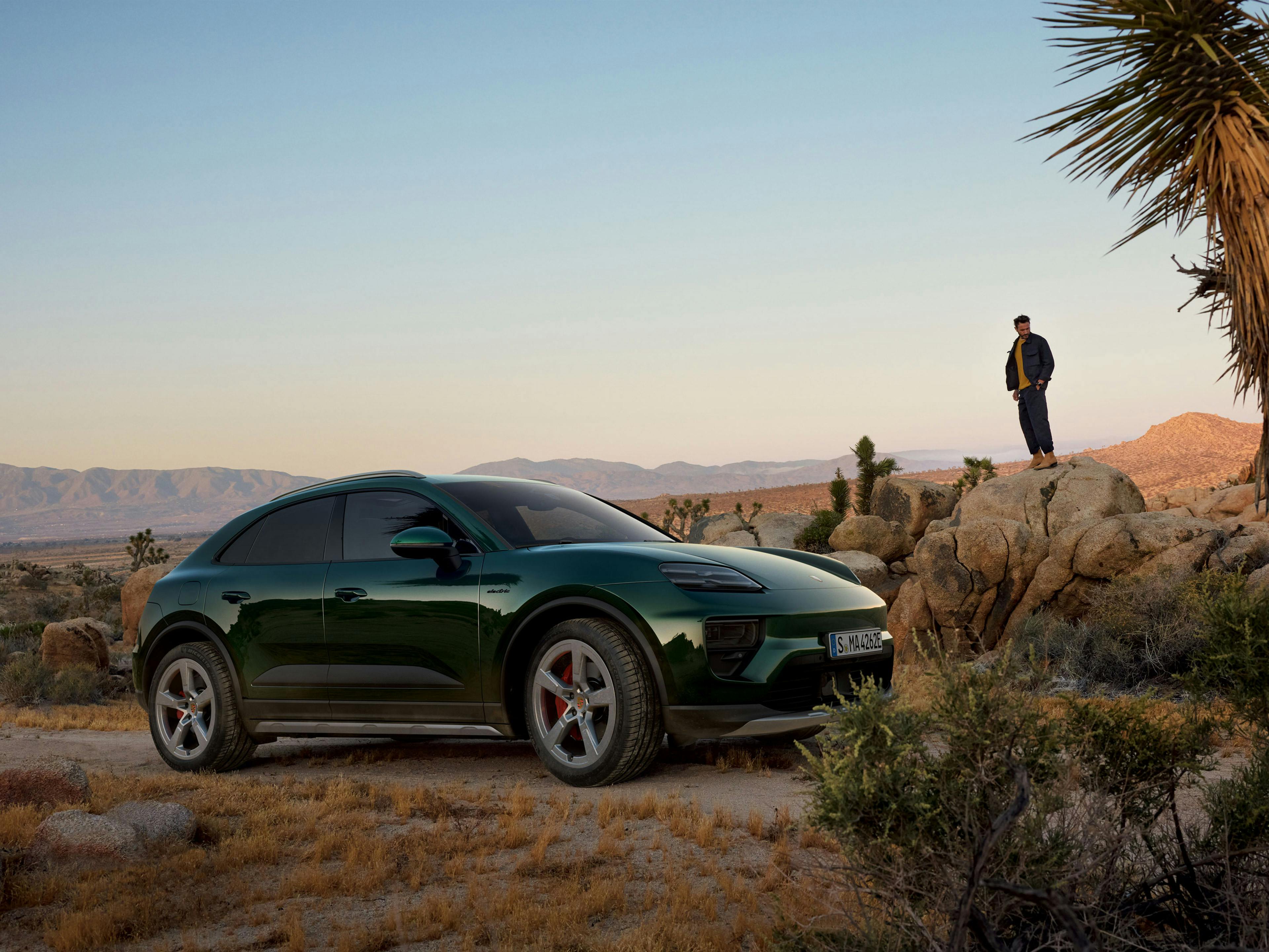 Green Porsche Macan 4S electric parked in the desert with a man standing on rock with a mountainous backdrop.
