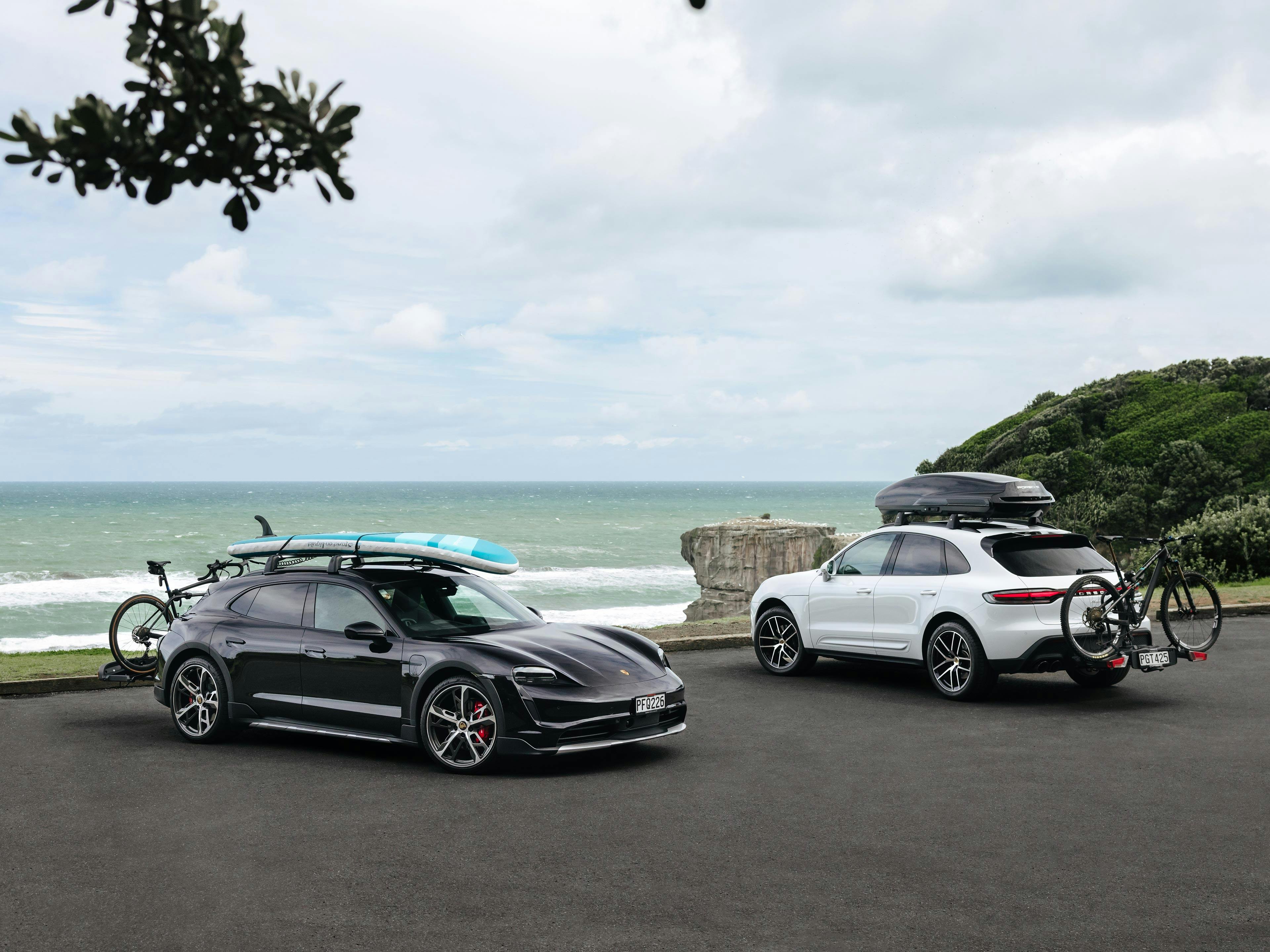 Two Porsche models parking near a coast. Both have a roof transport system with bikes & snowboards loaded.
