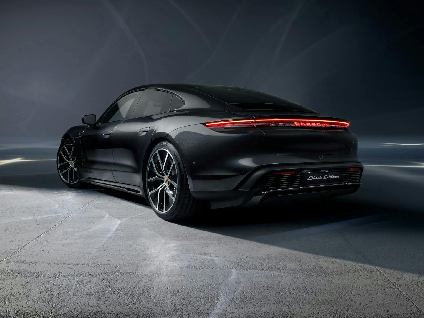Schwarzer Porsche Taycan in Heck‑Seitenansicht mit durchgehendem LED‑Leuchtenband vor dunklem Studiohintergrund.