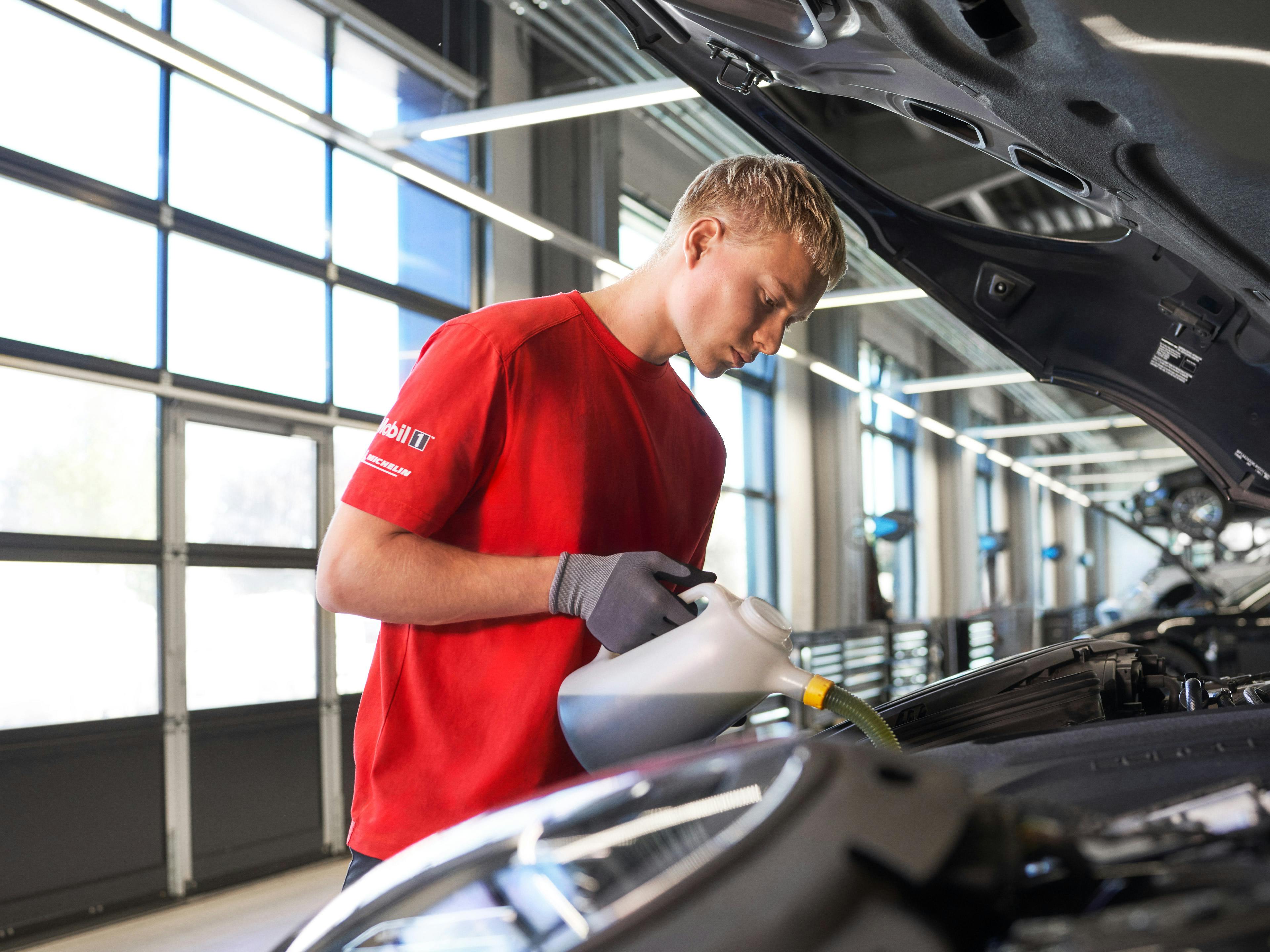 Ein junger Werkstattmitarbeiter in rotem T-Shirt und Handschuhen füllt Motoröl in ein Auto. Er arbeitet konzentriert in einer hellen Werkstatt mit großen Fenstern.