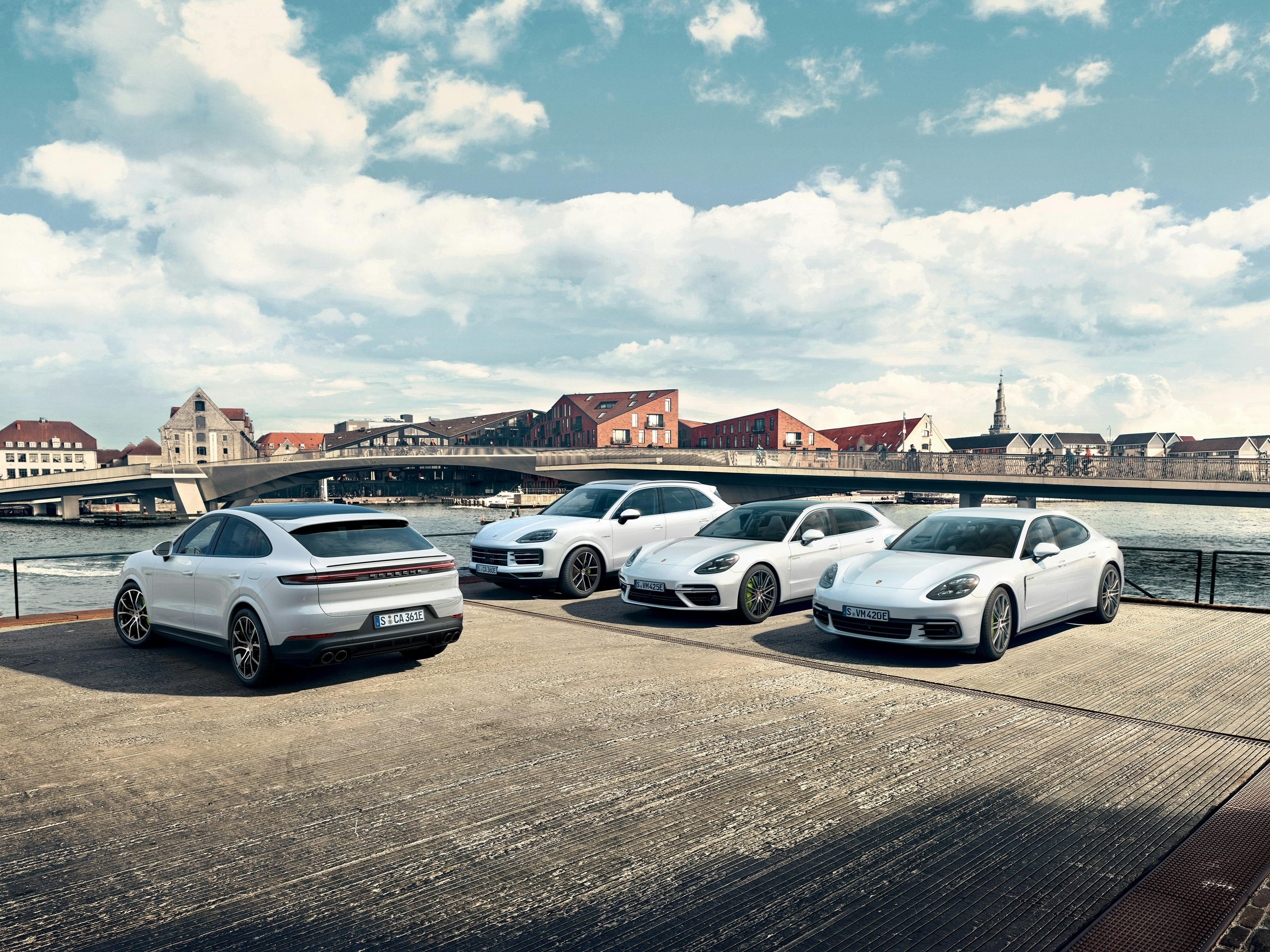 Vier weiße Porsche-Modelle (Cayenne und Panamera) stehen nebeneinander vor einer Brücke in einer Stadt mit historischem Flair.