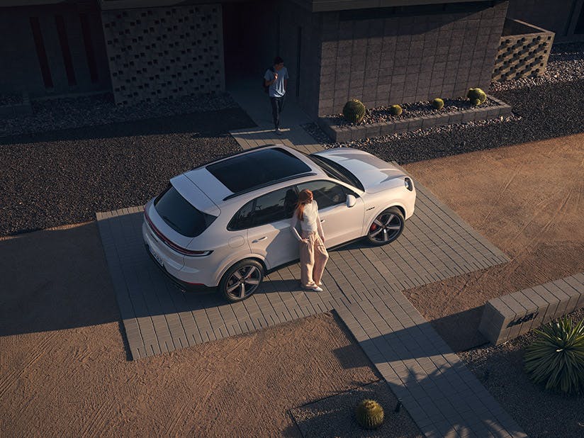 Luftaufnahme eines modernen Wohnhauses. Ein weißer Porsche Cayenne steht in der gepflasterten Einfahrt, eine Person neben der geöffneten Fahrertür.