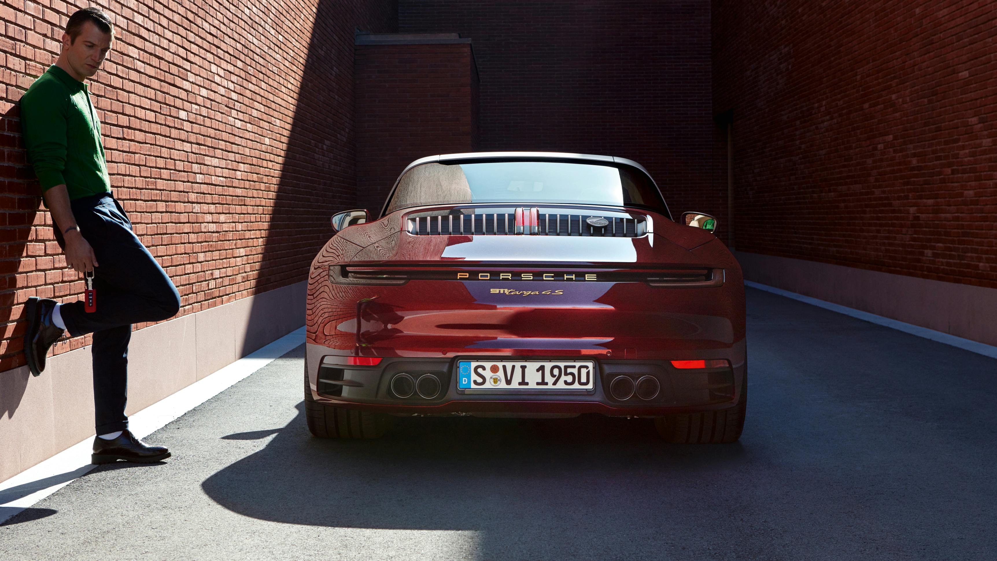 Porsche 911 Targa 4S Heritage Design Edition in Cherry Metallic. View of rear end. A man is leaning against the wall next to it.