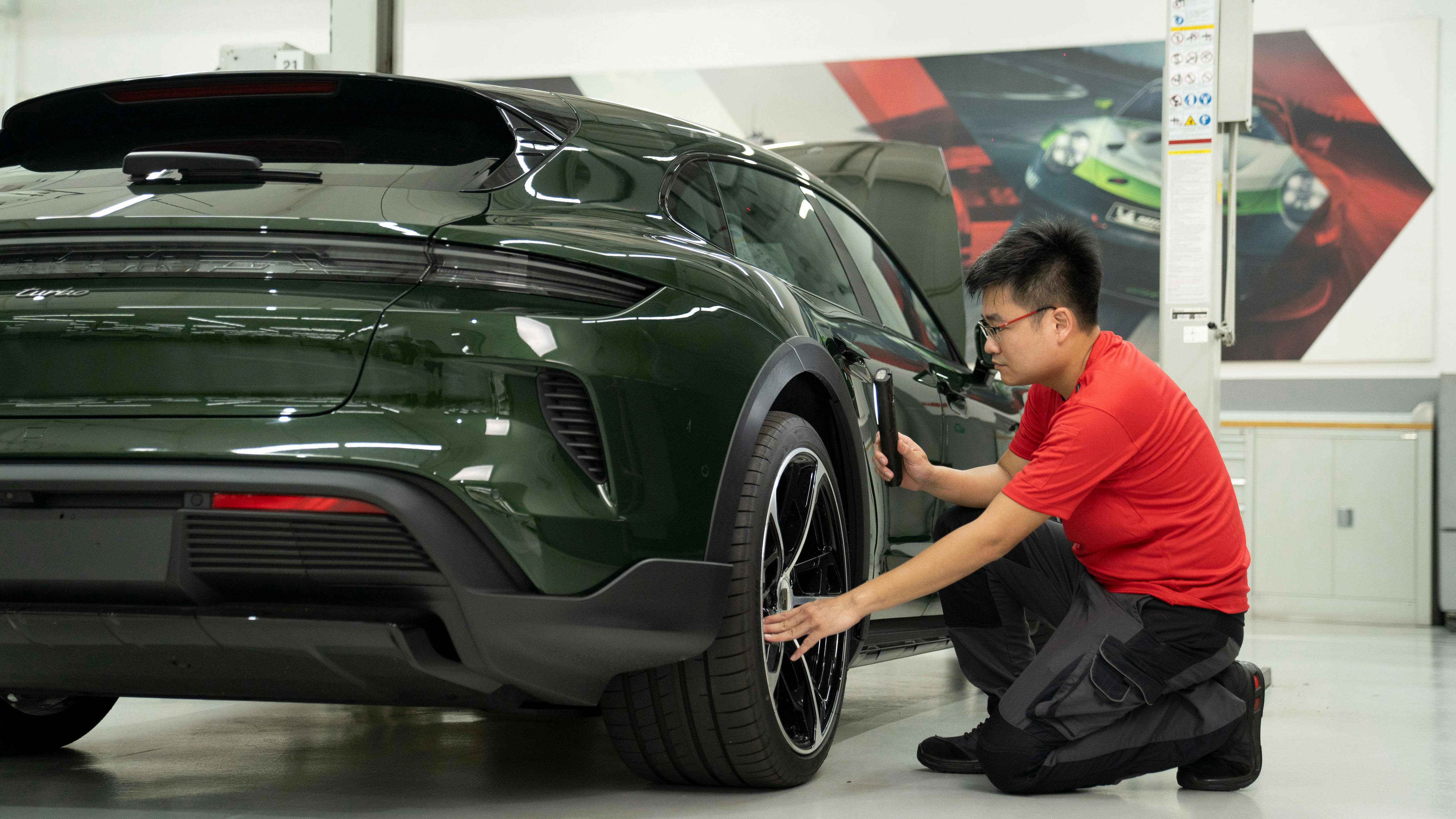 A technician in a red shirt inspecting the rear wheel of a dark green Porsche in a clean, brightly lit workshop.