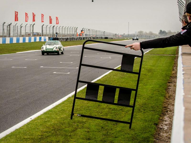 The ‘Project 70’ 911 races along the racetrack Donington Park Circuit. A man holds a sign with the lap information.