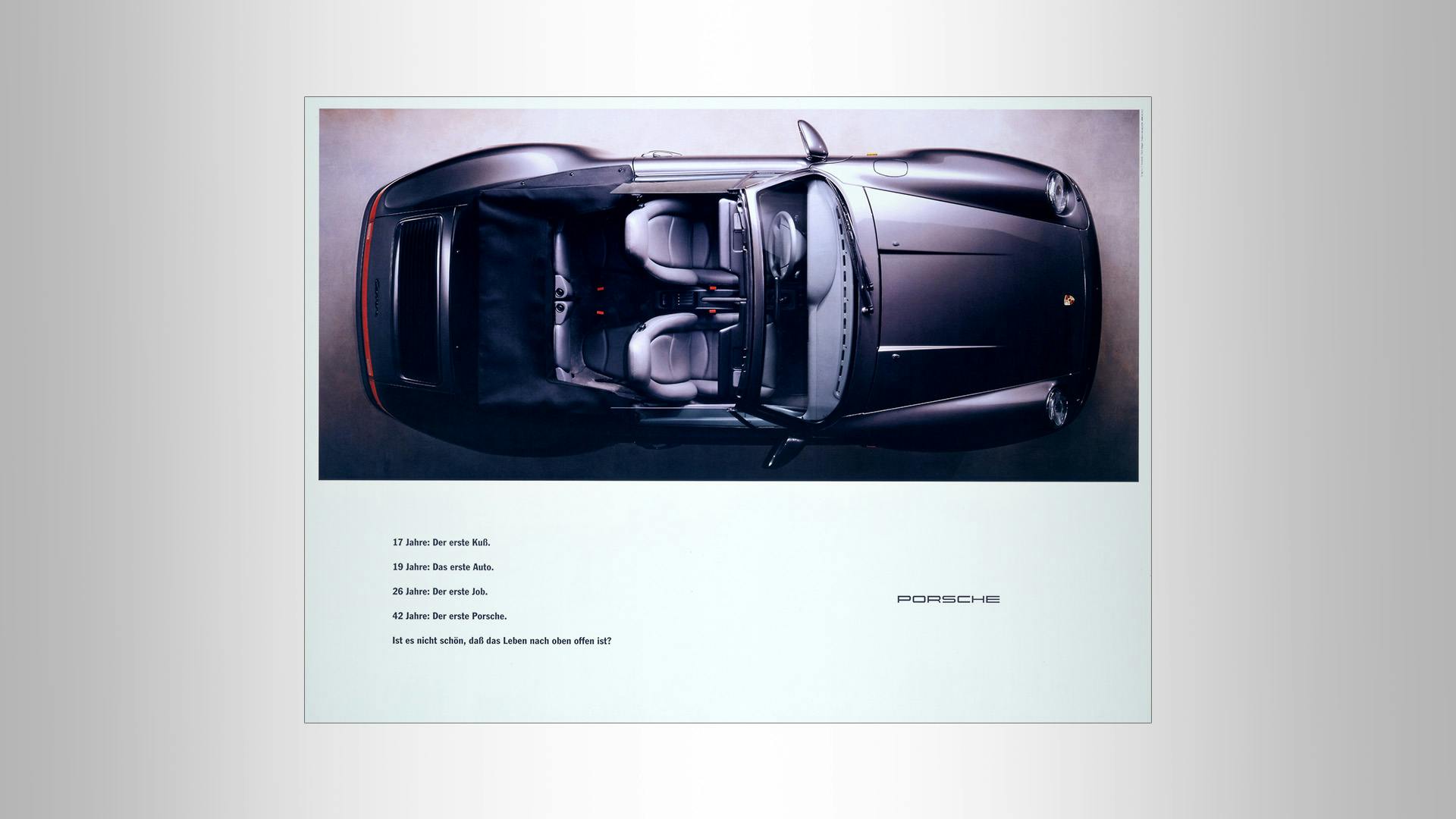 Porsche advertising poster with a black Porsche from above and the slogan: 17 Jahre: Der erste Kuß. 19 Jahre: Das erste Auto. 26 Jahre: Der erste Job. 42 Jahre: Der erste Porsche. Ist es nicht schön, daß das Leben nach oben offen ist?