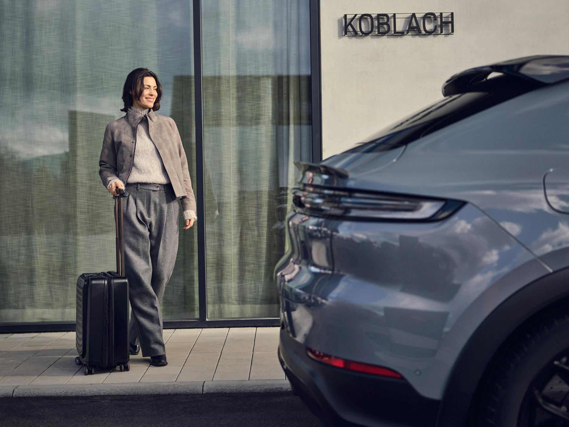 A woman with a trolley suitcase standing on the side of the road next to a Porsche Macan 4.