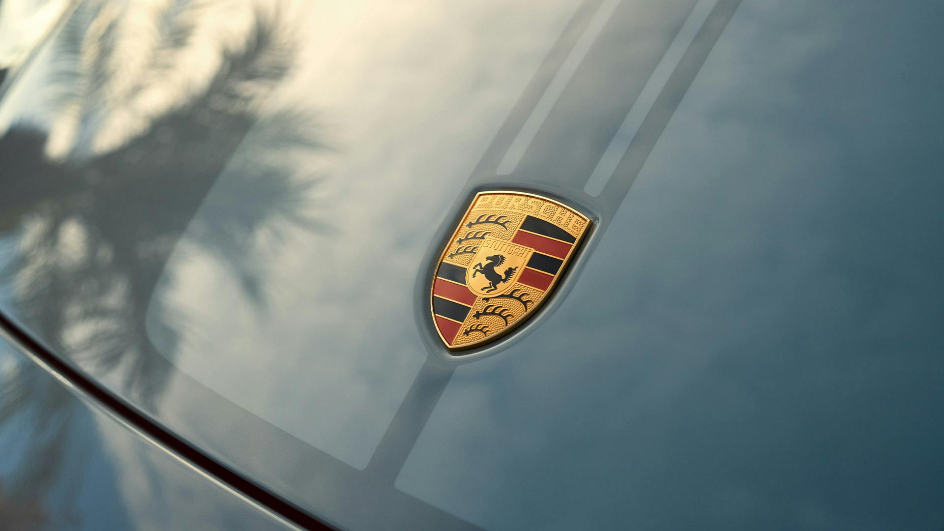 Ein Porsche-Emblem geprägt auf einer blauen Motorhaube, überschattet von den Silhouetten der Bäume.