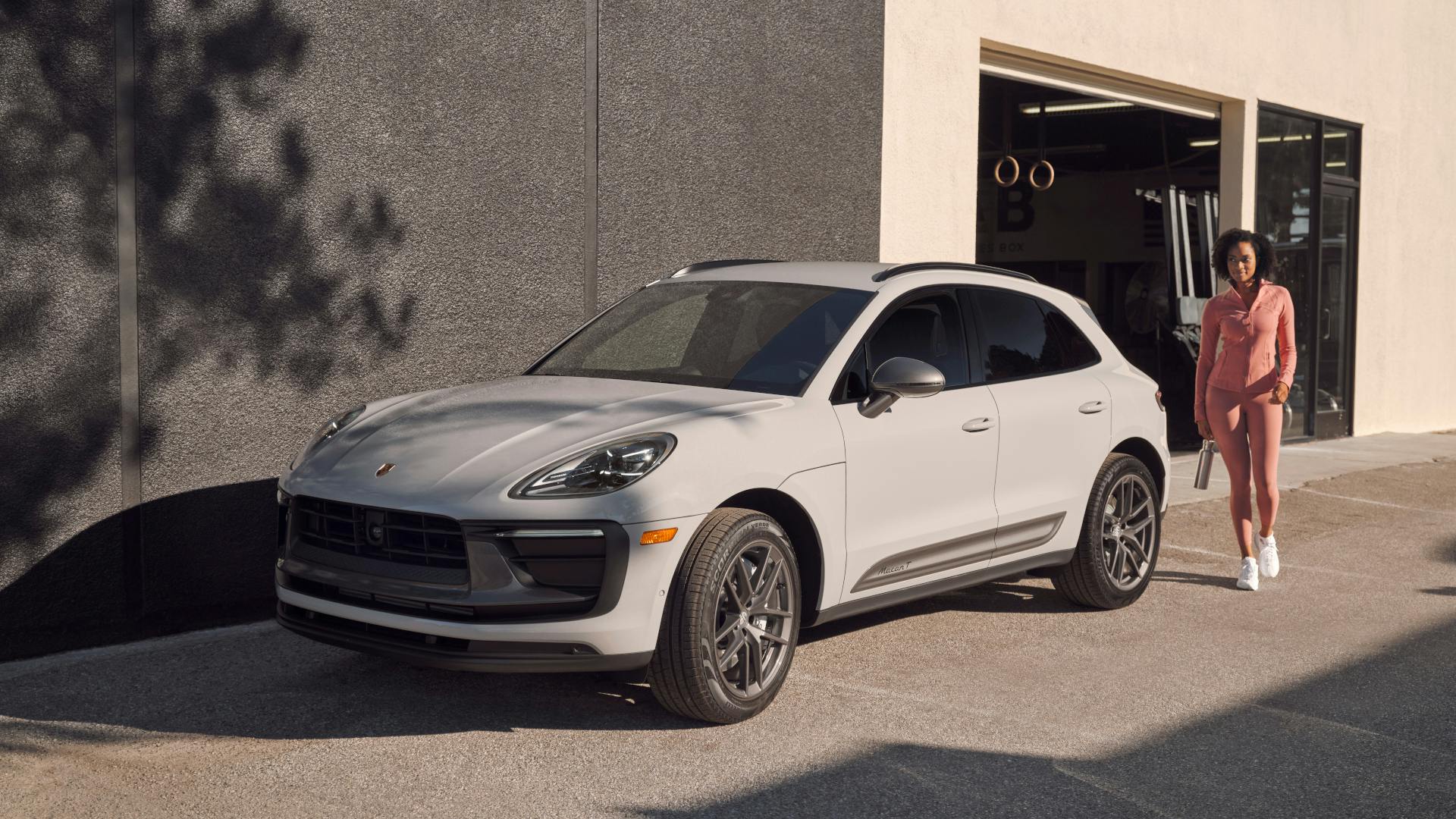 A light grey Porsche Macan T parked in front of a modern building, with a person standing nearby in casual athletic clothing on a sunny day.