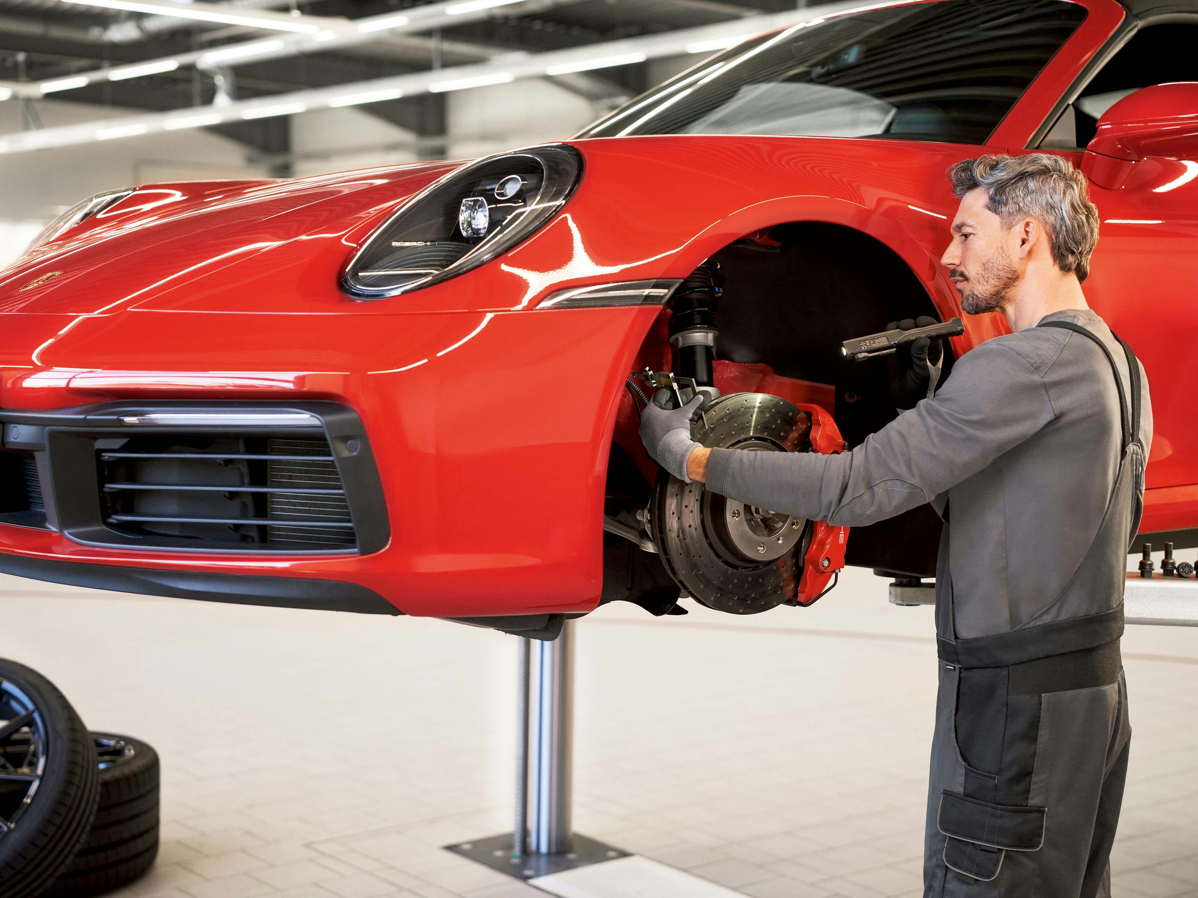 Een monteur is bezig met de remmen van een Porsche. De auto is deels opgetild. De monteur leunt over de band en kijkt geconcentreerd naar het werk dat hij doet.