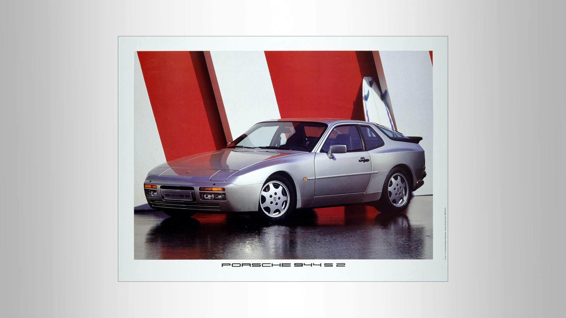 Advertising silver Porsche 944 S 2 standing in front of a red/white wall.