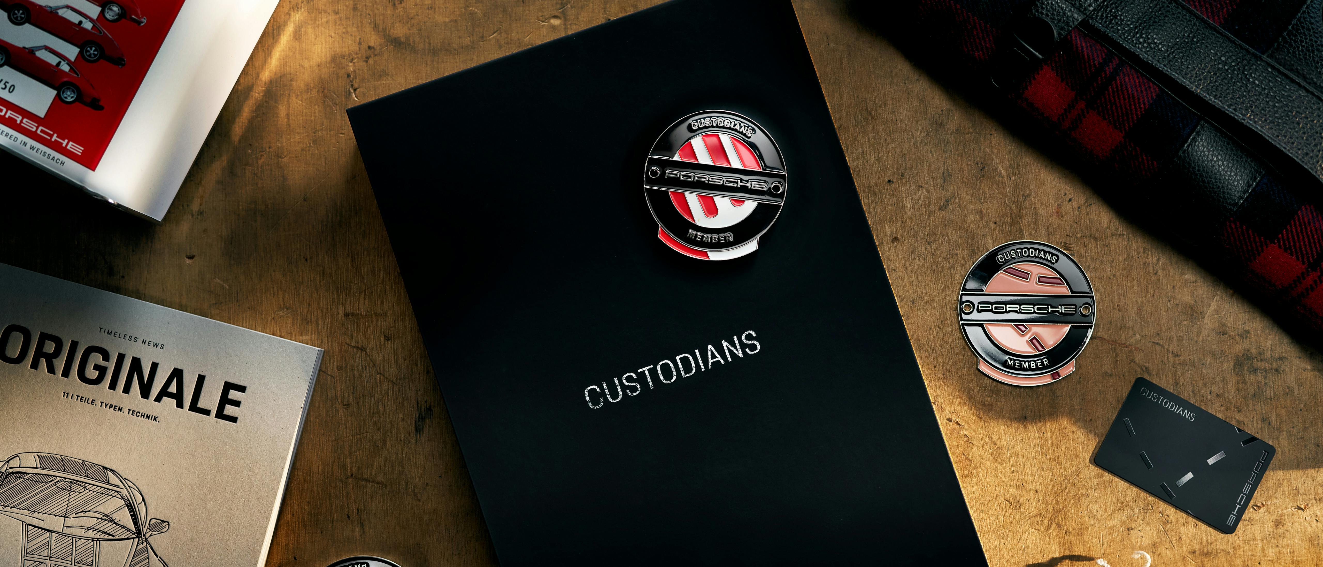 A top-down view of several Porsche-branded collector items arranged on a rustic wooden table. In the centre is a large, matte black book titled "CUSTODIANS" with the word "PORSCHE" at the bottom. Around it are three circular metallic badges featuring the Porsche logo and the word "MEMBER," a small black membership card, a grey "ORIGINALE" booklet with a car sketch, a red and black plaid bag, and a vintage-style white tin.