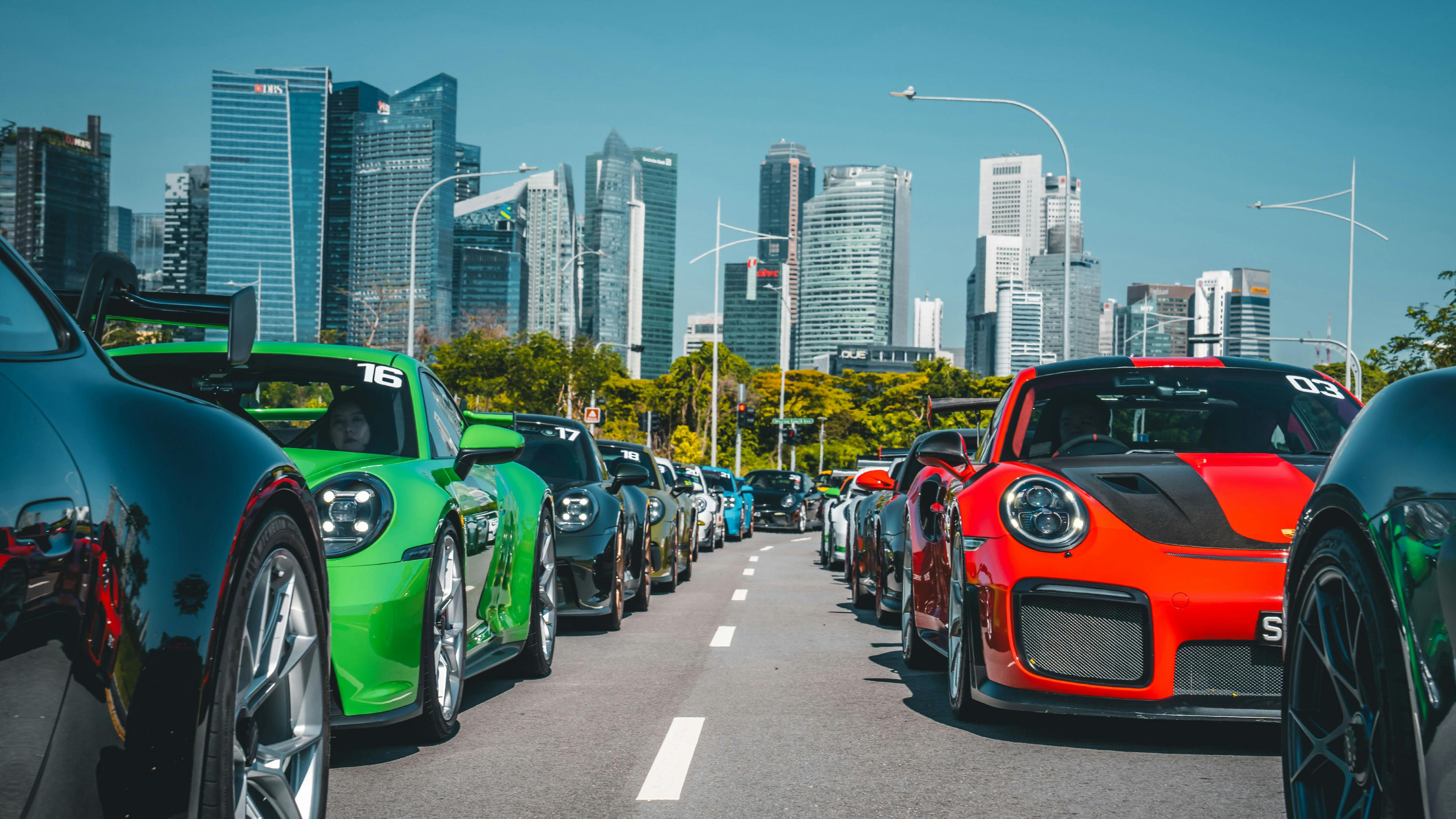 Two rows of Porsche cars of various colours set against the Singapore building skyline in the day