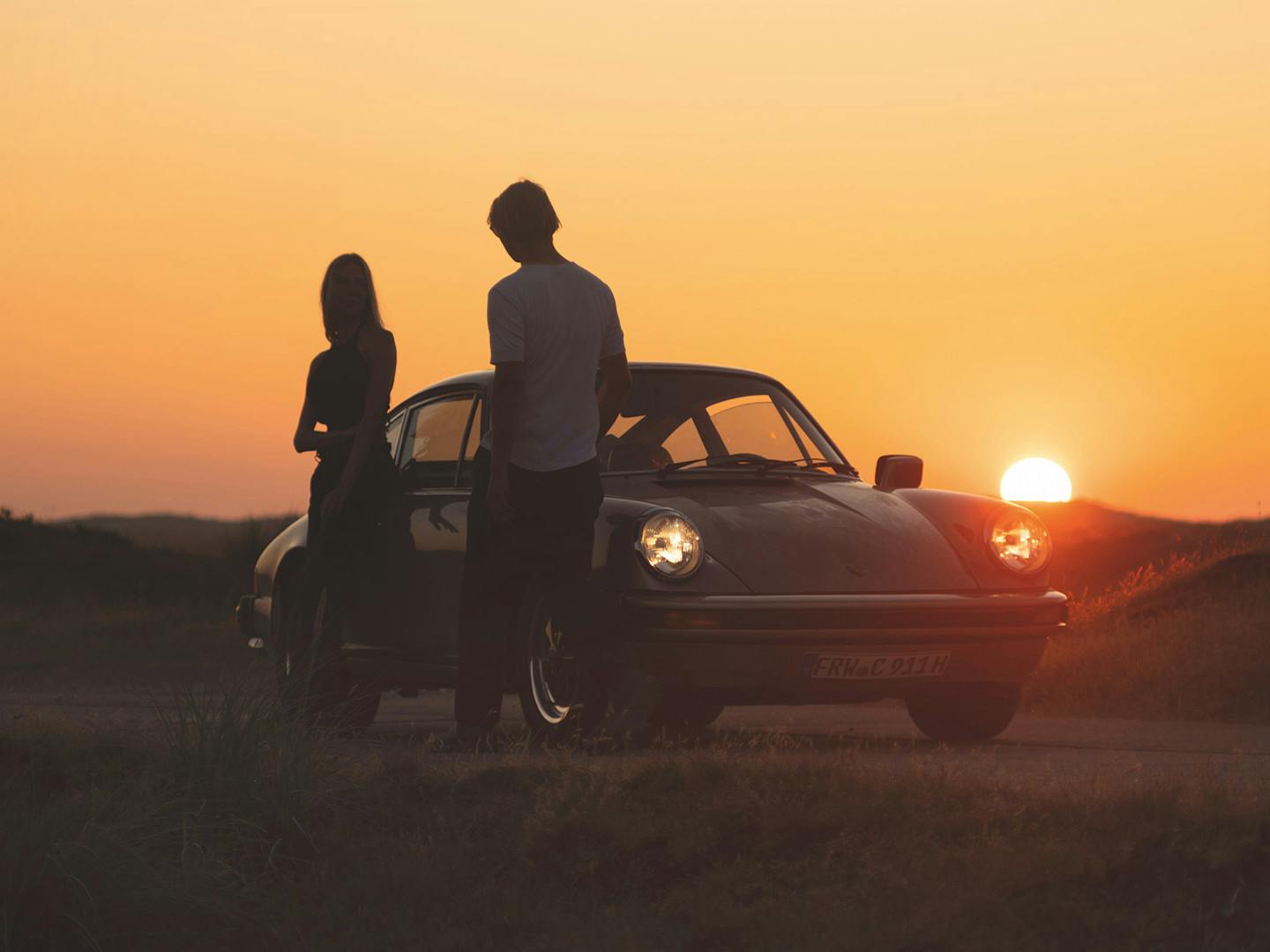 Two persons standing next to a Porsche classic car during sundown.