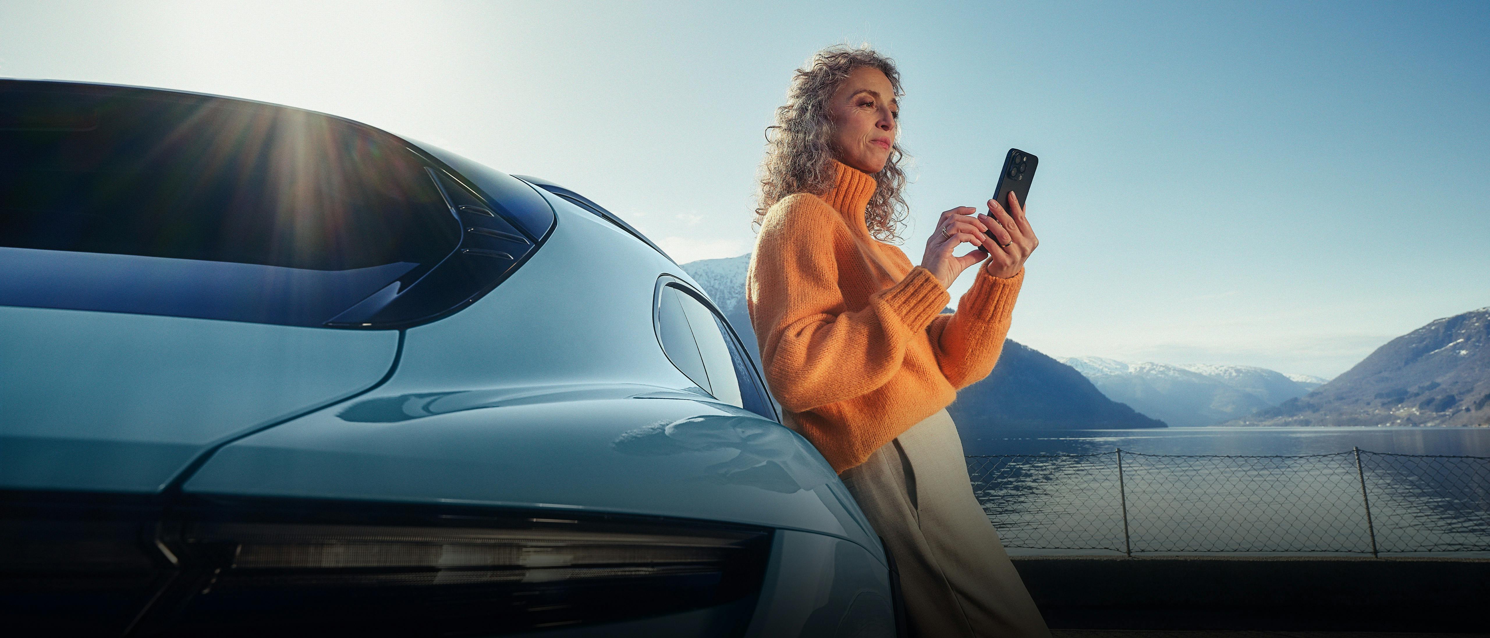 A woman looking at her phone while leaning against a parked light blue Porsche Taycan.