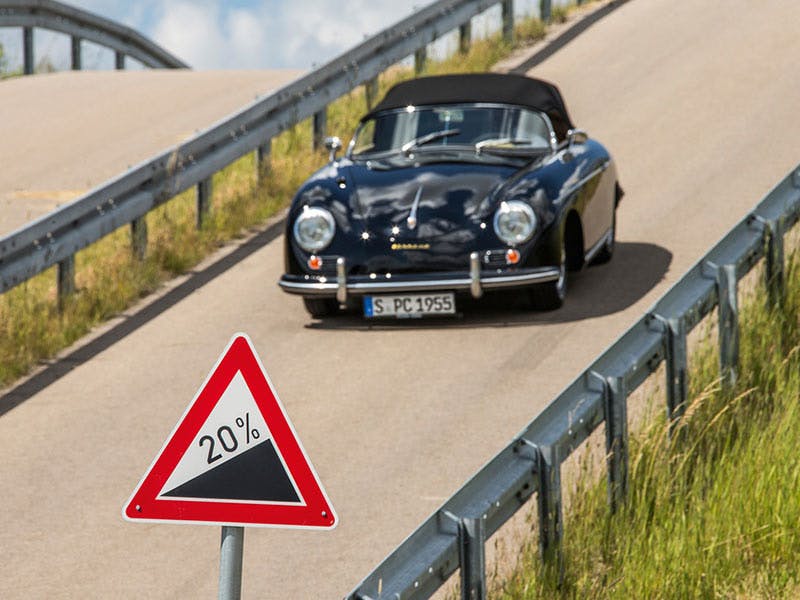 Image: Black Porsche 356 on road with a 20% incline, 20% road sign, crash barriers.