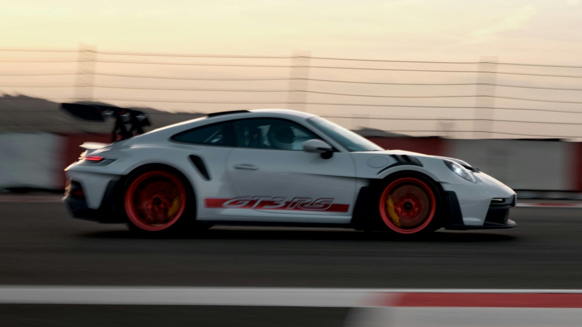 A white Porsche 911 GT3 RS drives at high speed on a racetrack at sunset.