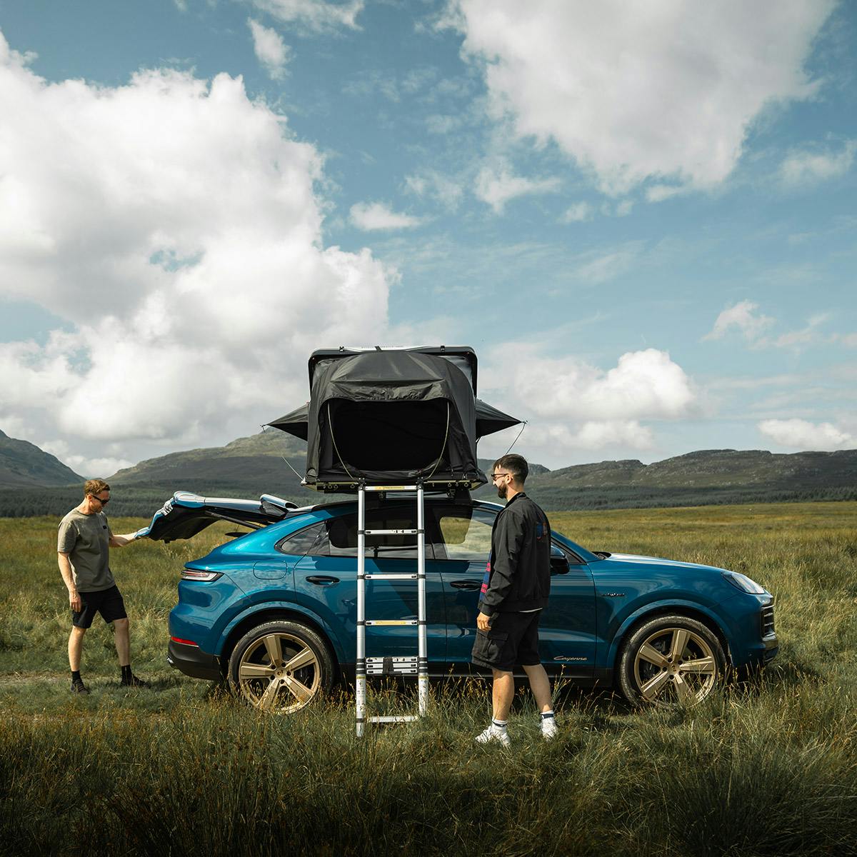 Two men standing next to a blue car with a tent on top in the countryside.​