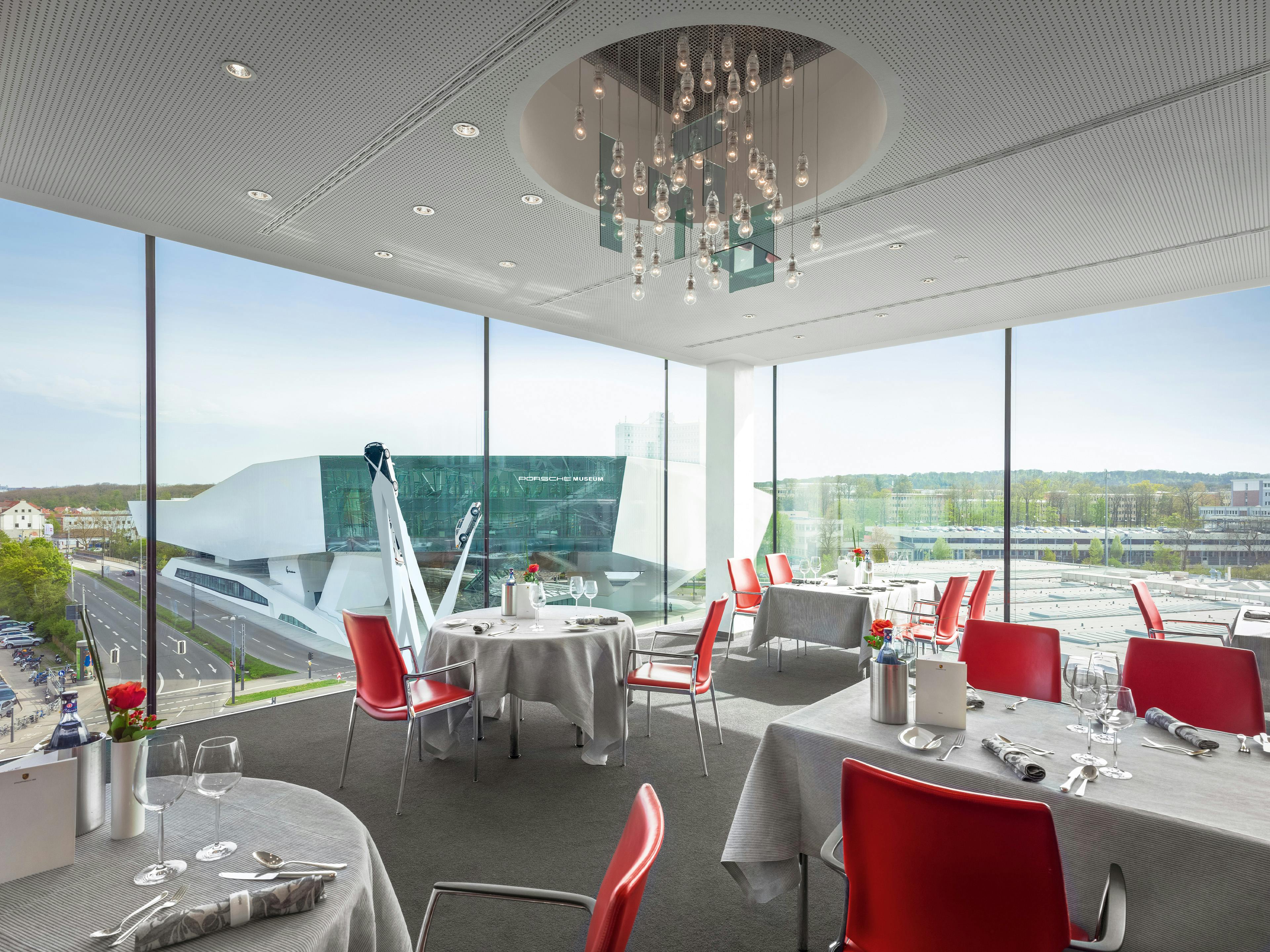 Contemporary dining room at Restaurant Christophorus overlooking the Porsche Museum, featuring red accent chairs and floor-to-ceiling windows.