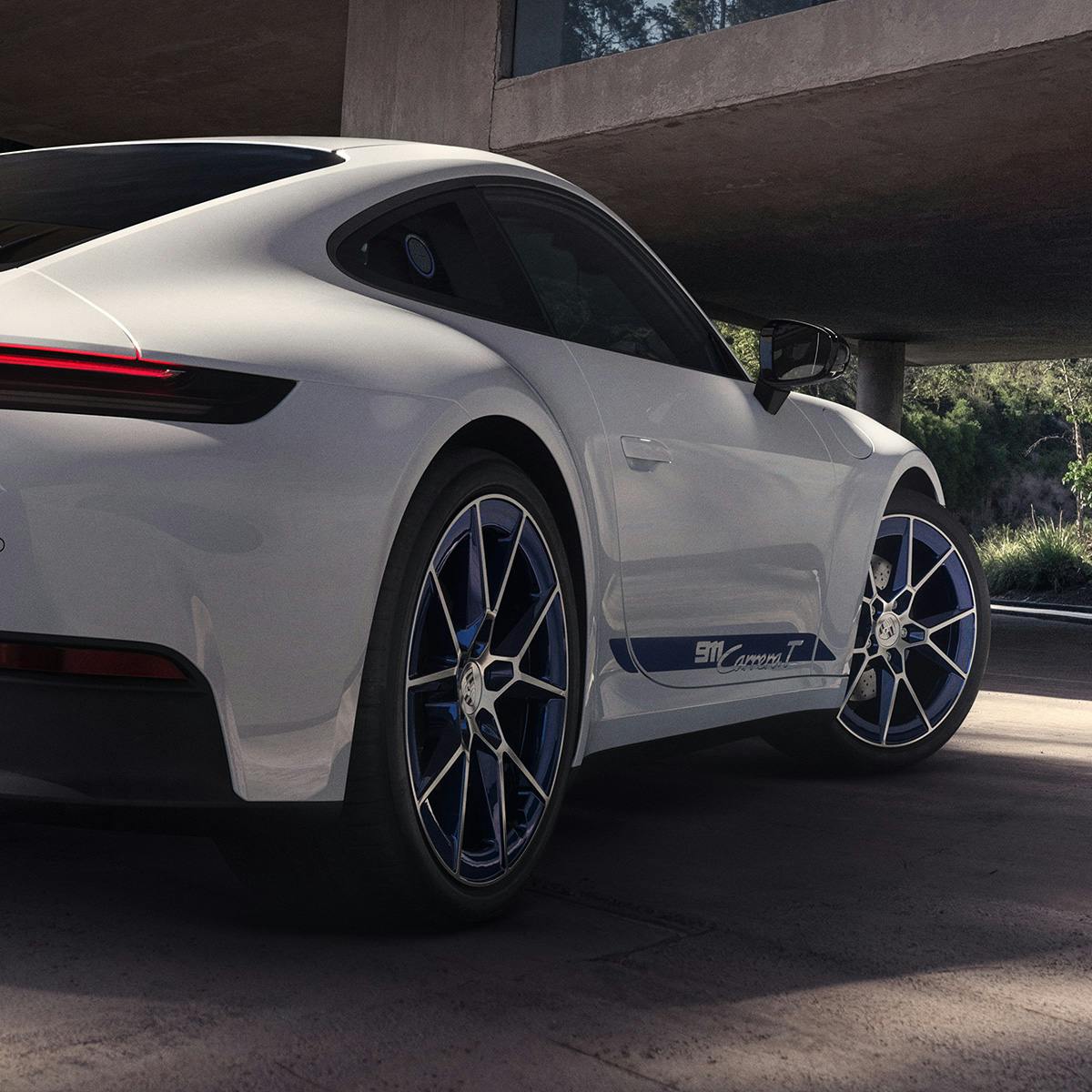 A white Porsche 911 Carrera T is shown from the rear three-quarter angle, parked under a modern concrete structure. The car features blue accents on the wheels and side.