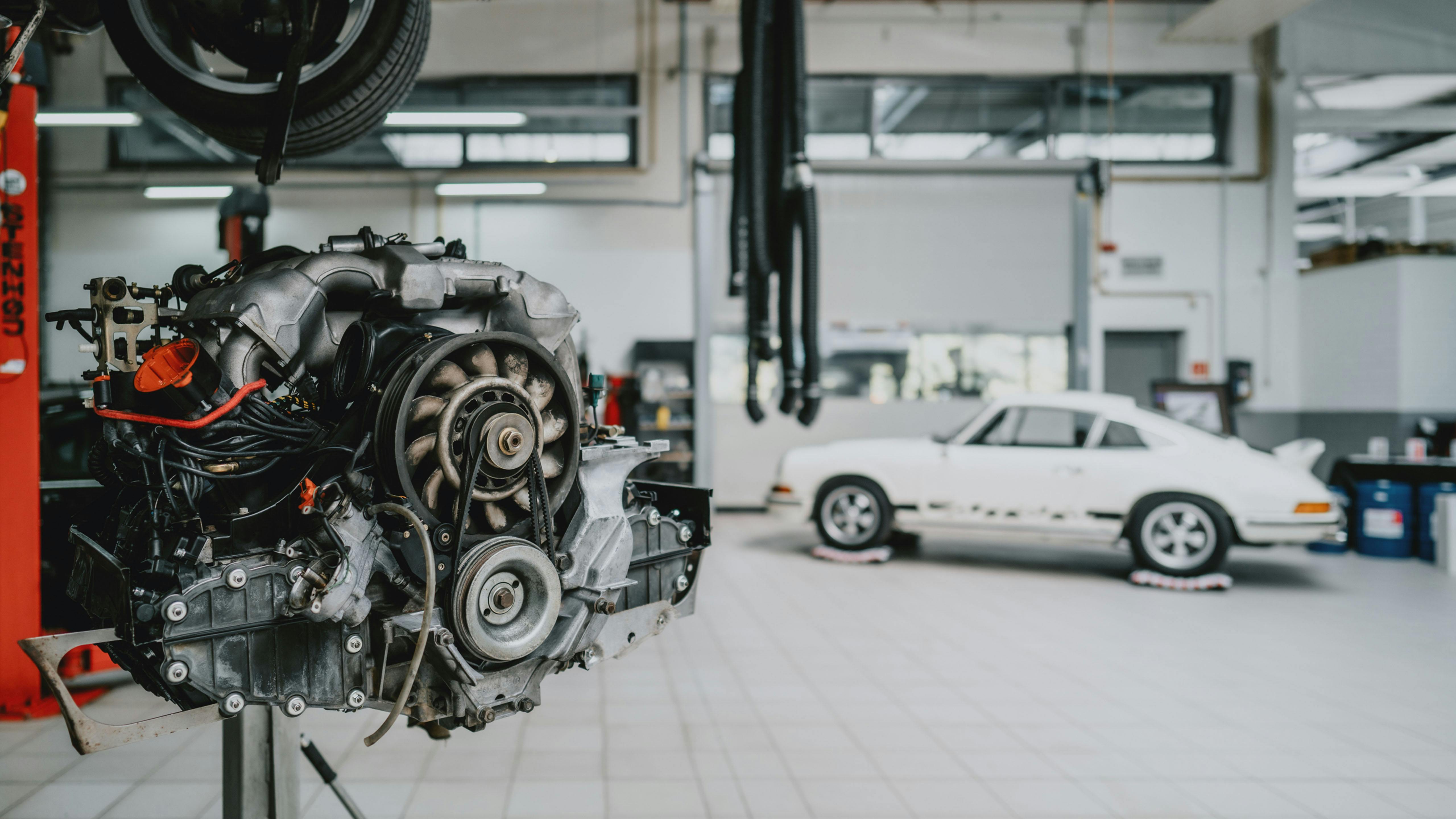 Rapproché d'un moteur avec une voiture Porsche blanche floue en arrière-plan dans l'atelier du Porsche Zenter Lëtzebuerg.