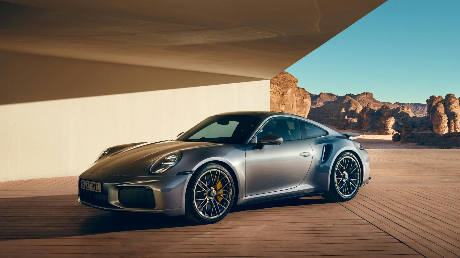 A silver Porsche 911 Turbo S parked under a modern architectural roof, with desert rock formations and a clear blue sky in the background.