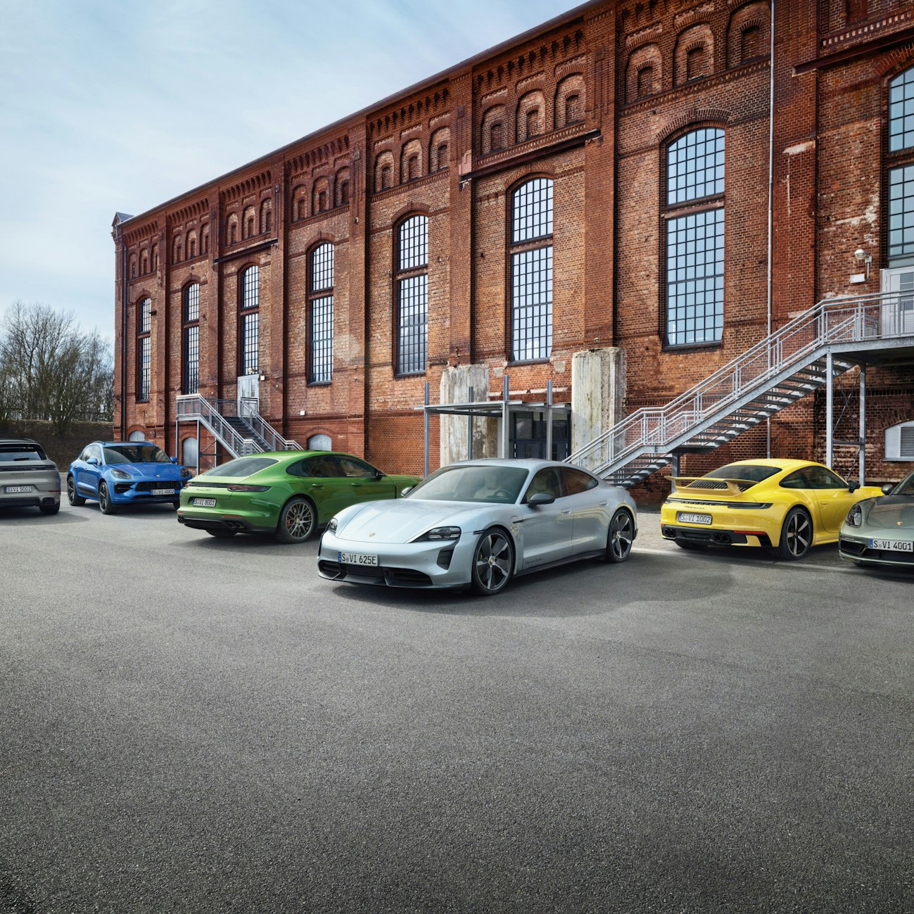 All six Porsche car models parked outside in front of a brick building.