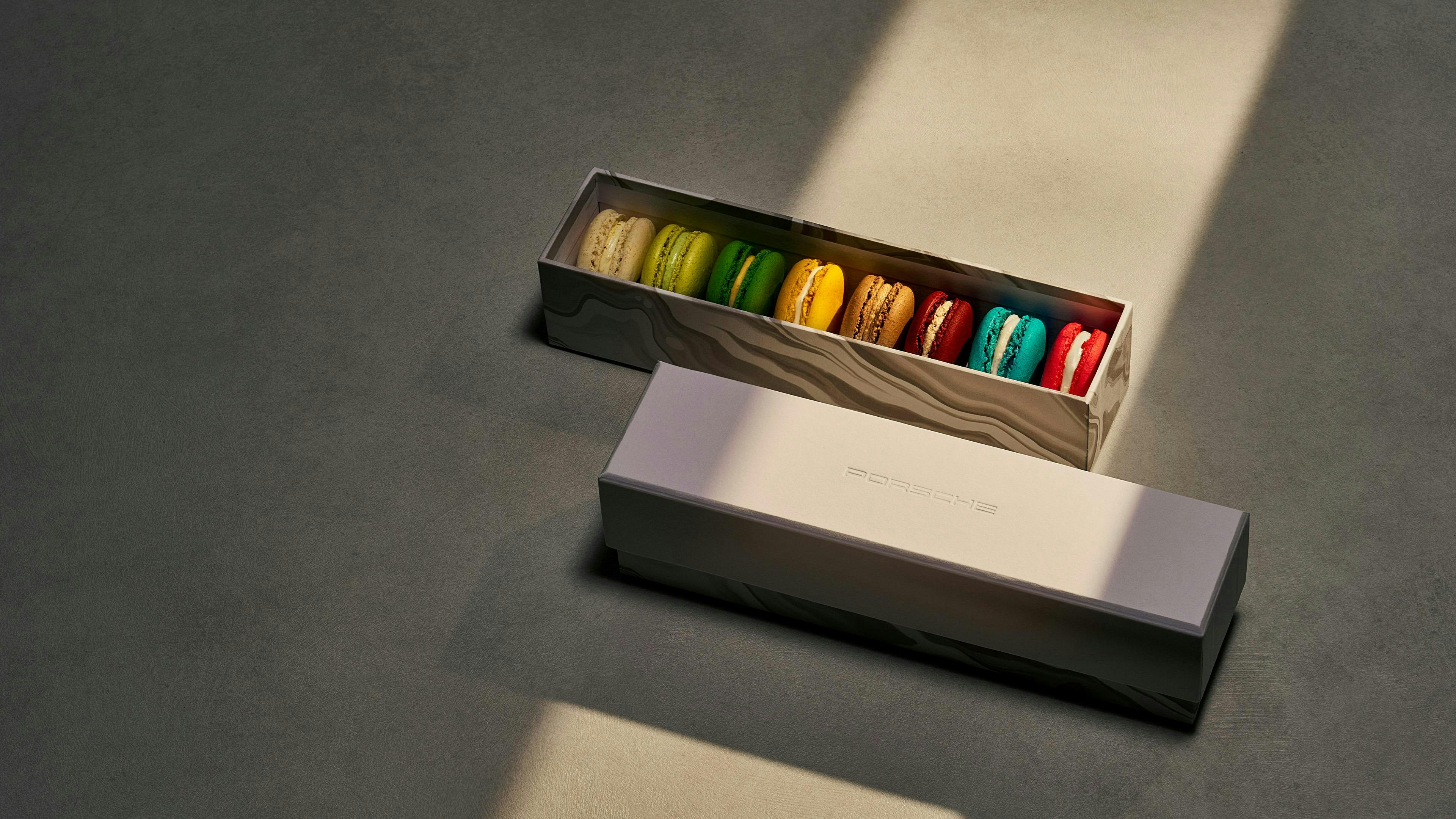 Box of colorful macarons in Porsche-branded packaging, arranged in a row with light highlighting the vibrant shades.