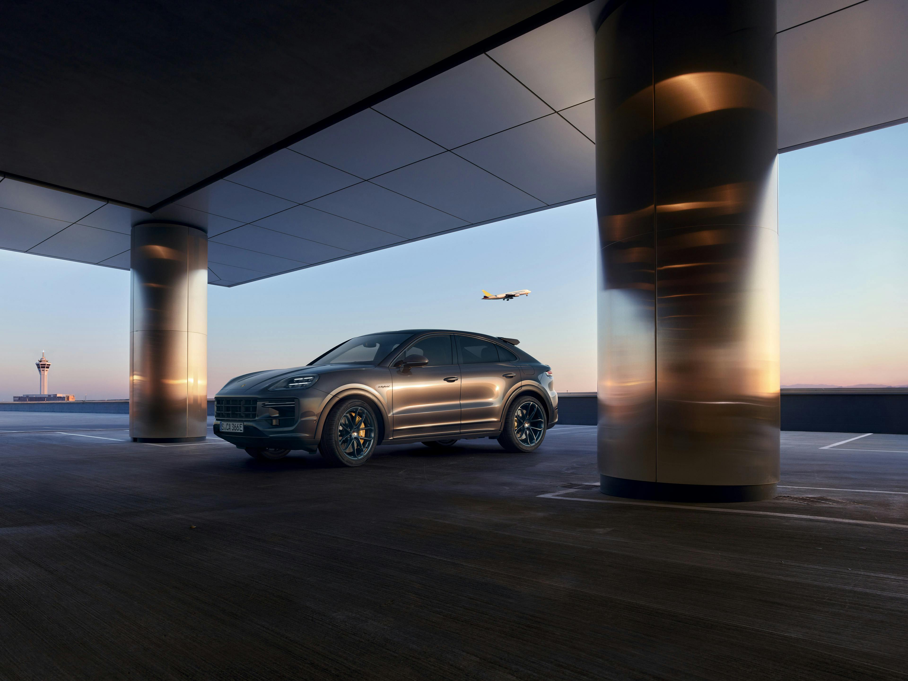 Ein dunkelgrauer SUV (Cayenne) mit sportlichem Design steht auf einem modernen Parkdeck bei Sonnenuntergang mit einem Flugzeug in der Luft. Die Szene ist von silber spiegelnden Säulen und einem klaren Himmel im Hintergrund umrahmt.