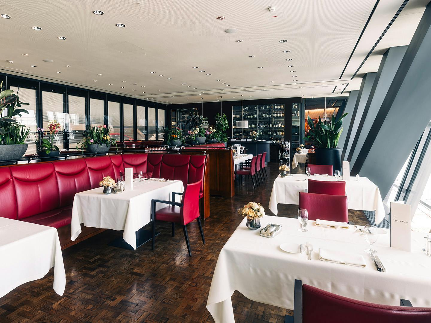 Restaurantbereich mit roten Lederbänken, weiß gedeckten Tischen und großen Pflanzen vor einer Glasfassade.