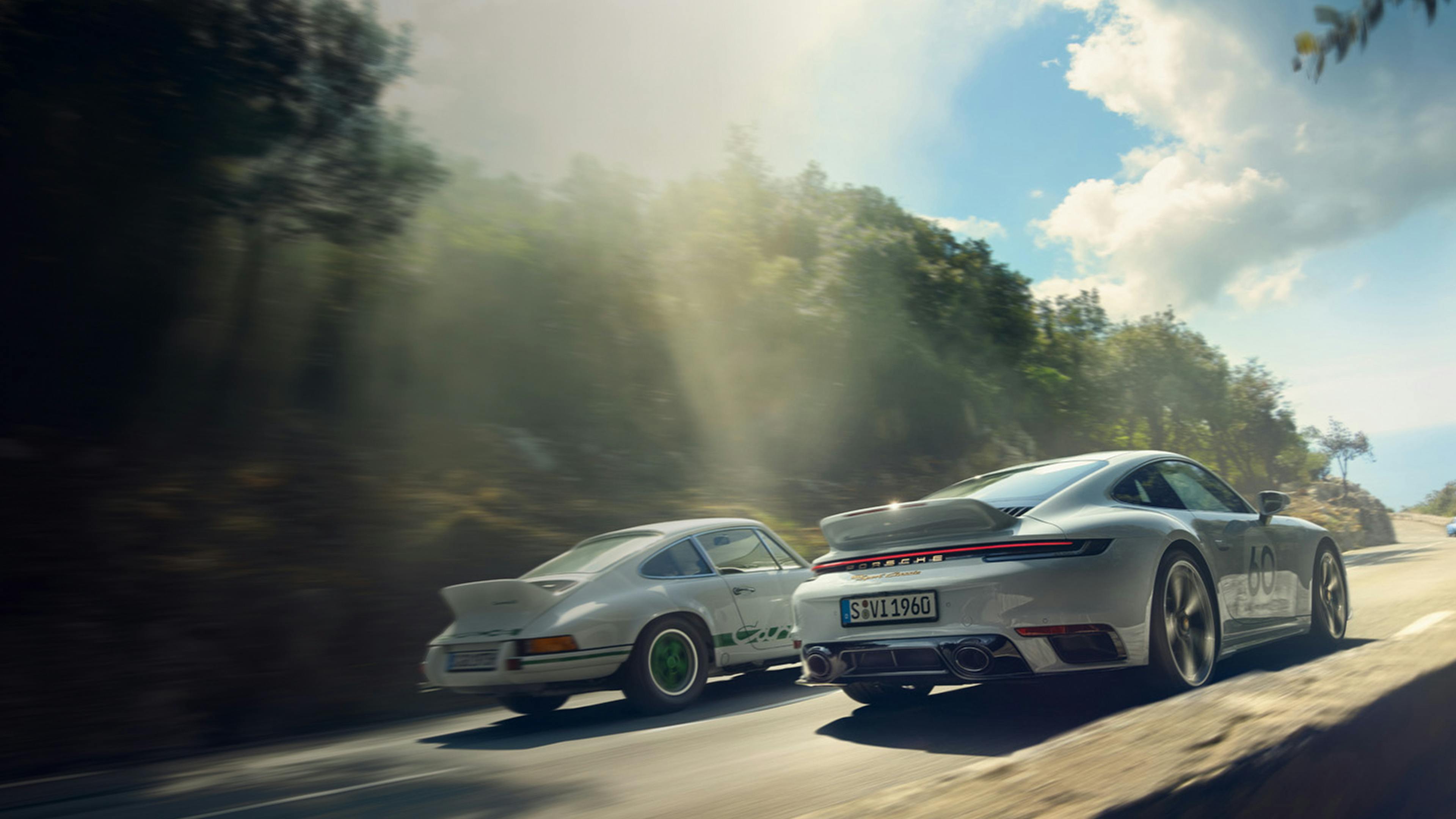 A Porsche 911 Sport Classic in Sport Grey Metallic driving along a coastal road next to a classic Porsche 911. View of rear end.