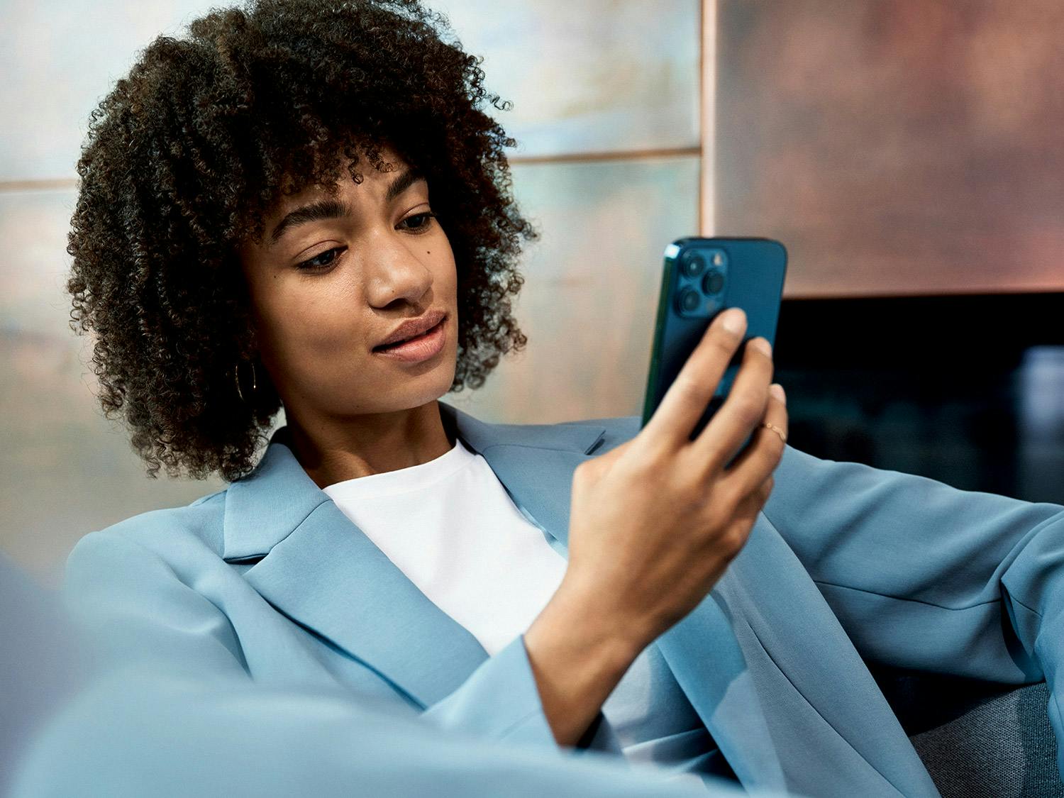 Une jeune femme aux cheveux noirs bouclés, vêtue d'un blazer bleu clair , regarde son smartphone.