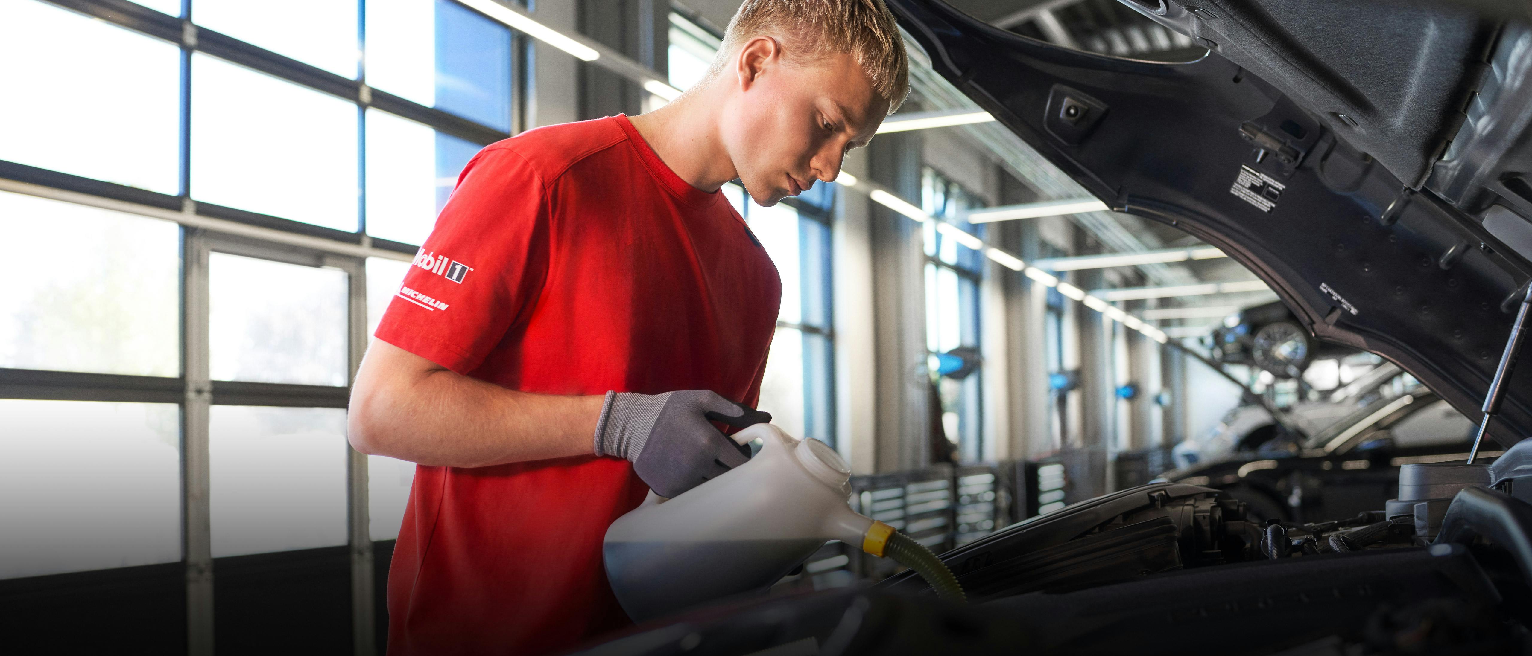 Un technicien automobile portant un t-shirt rouge remplissant l’huile moteur d’une Porsche dans un atelier de service.