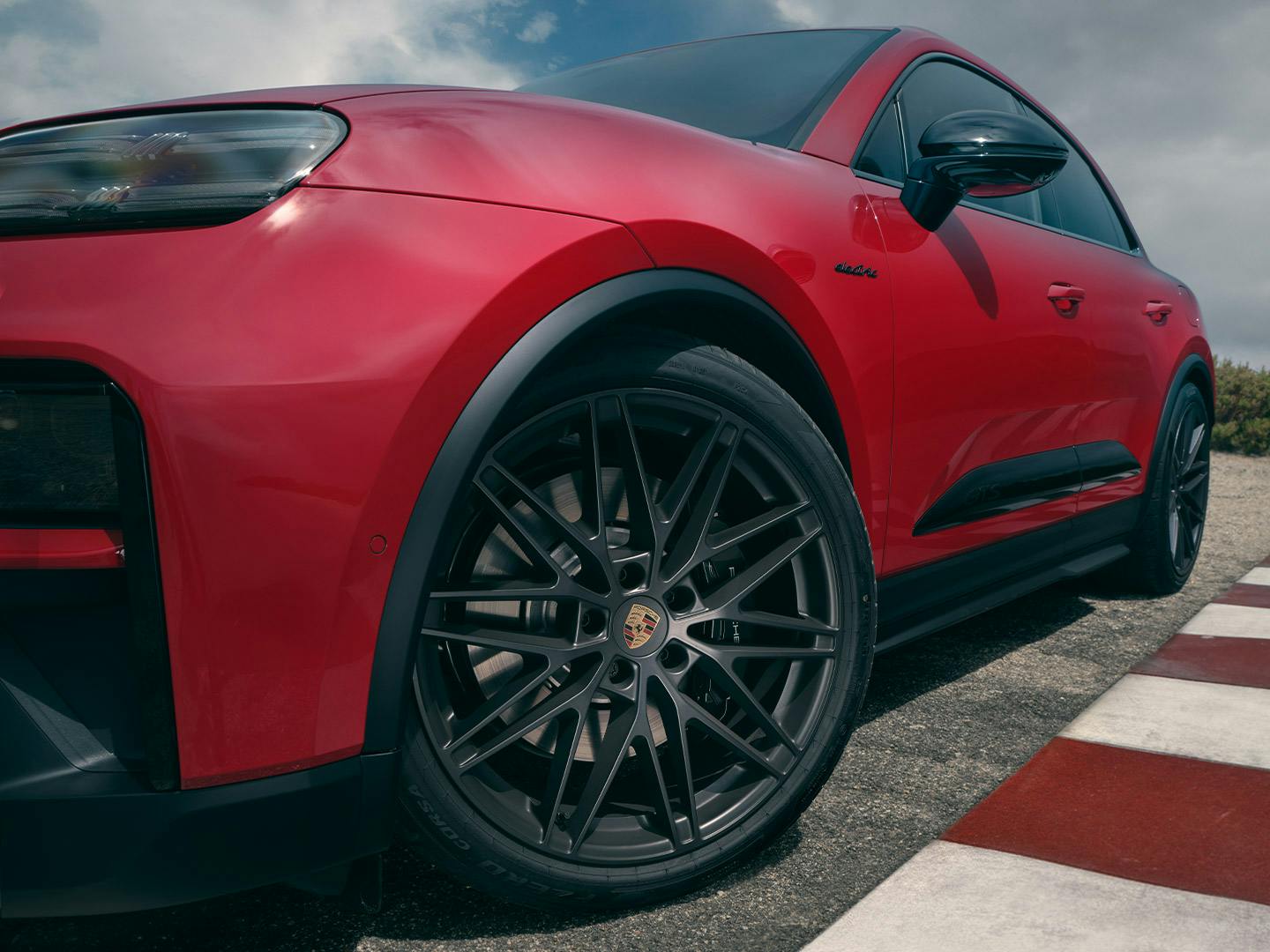 Nahaufnahme eines roten Porsche Macan. Im Vordergrund das vordere Rad mit dunkler Felge und sichtbarem Porsche‑Emblem auf einer Rennstrecke.