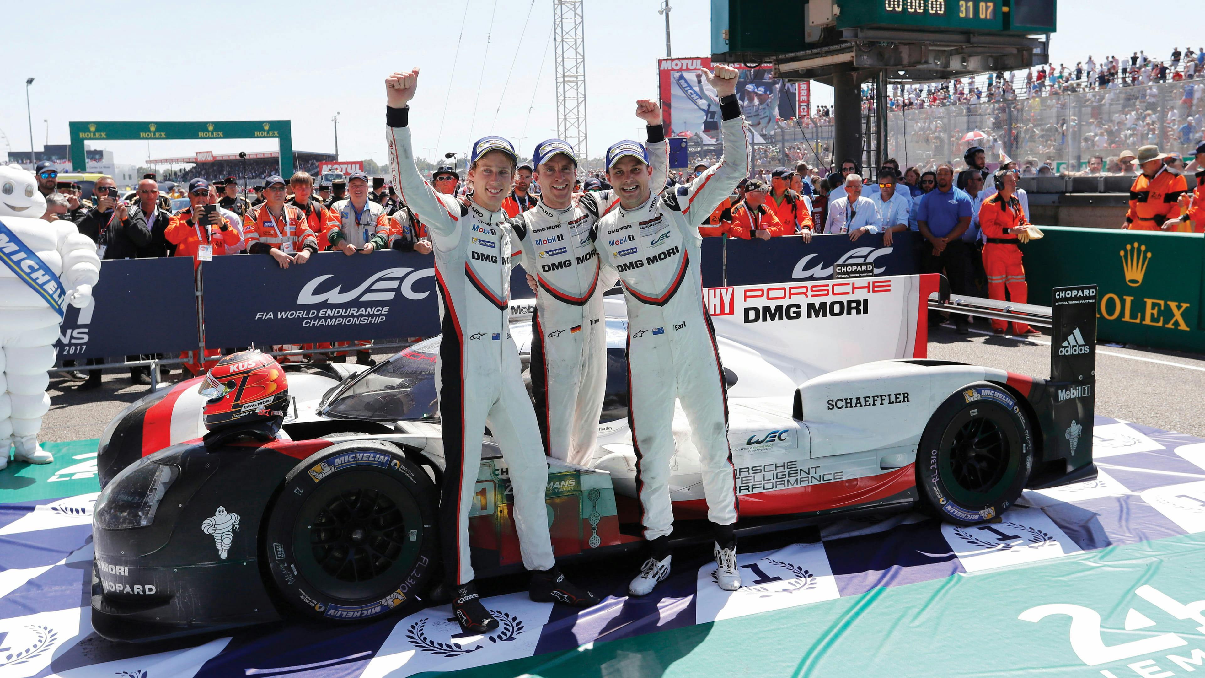 Three racing drivers standing beside a race car on a track, raising their arms, with spectators in the background.