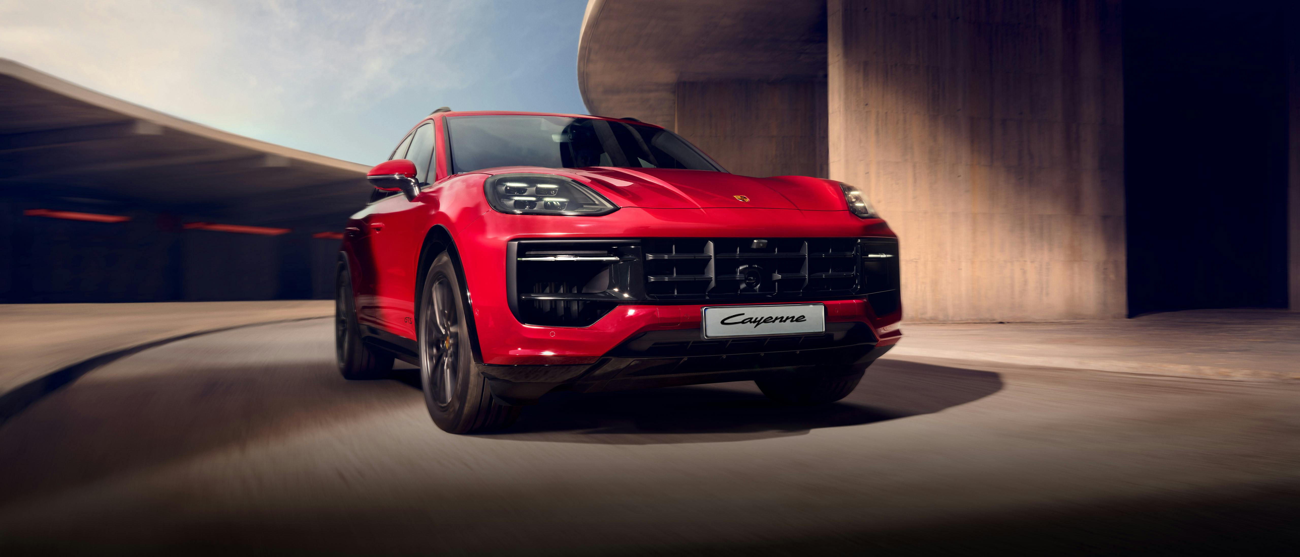 A red Porsche Cayenne SUV is shown driving dynamically in an urban setting, highlighting its sporty design and powerful presence.