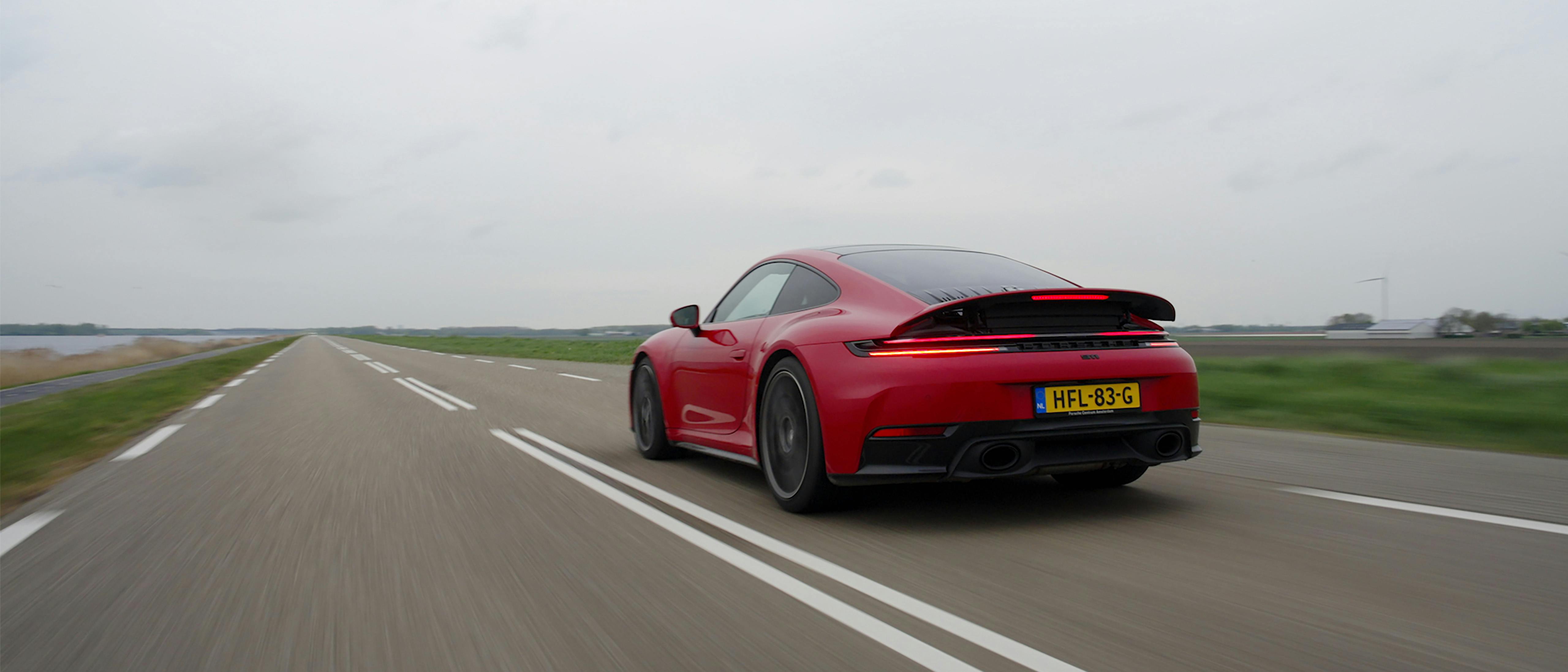 Achteraanzicht van een rode Porsche 911 (type 992) rijdend op rechte een polderweg. De actieve spoiler is uitgeklapt, de led-verlichting brandt en de dubbele sportuitlaten zijn zichtbaar onder het Nederlandse kenteken. Het lage standpunt accentueert de brede achterzijde en het iconische silhouet van de sportwagen.