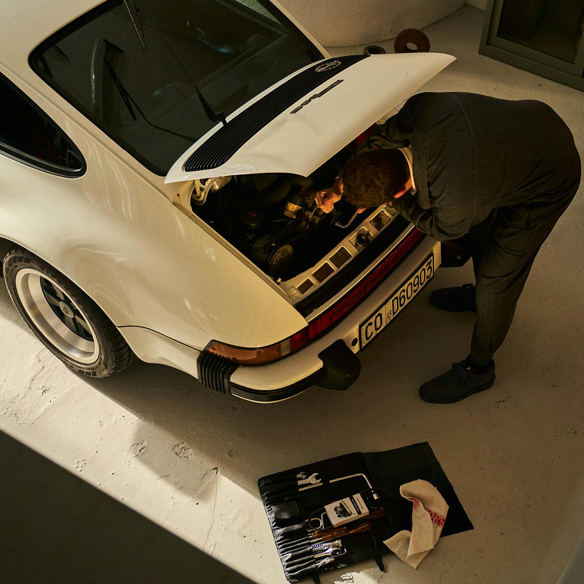 Una persona está inclinada sobre el compartimento del motor abierto de un Porsche 911 G blanco; junto a él hay una bolsa de herramientas abierta en el suelo.