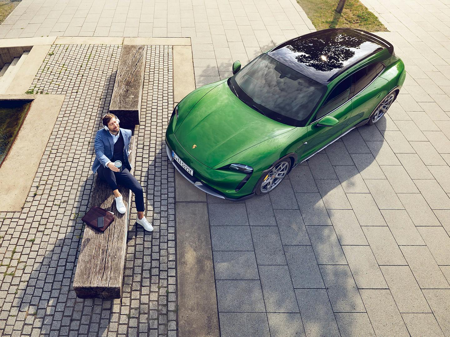 A bird's eye view of a Green Porsche Taycan in a paved car park next to a pond. A man with headphones is sitting in front of the car.