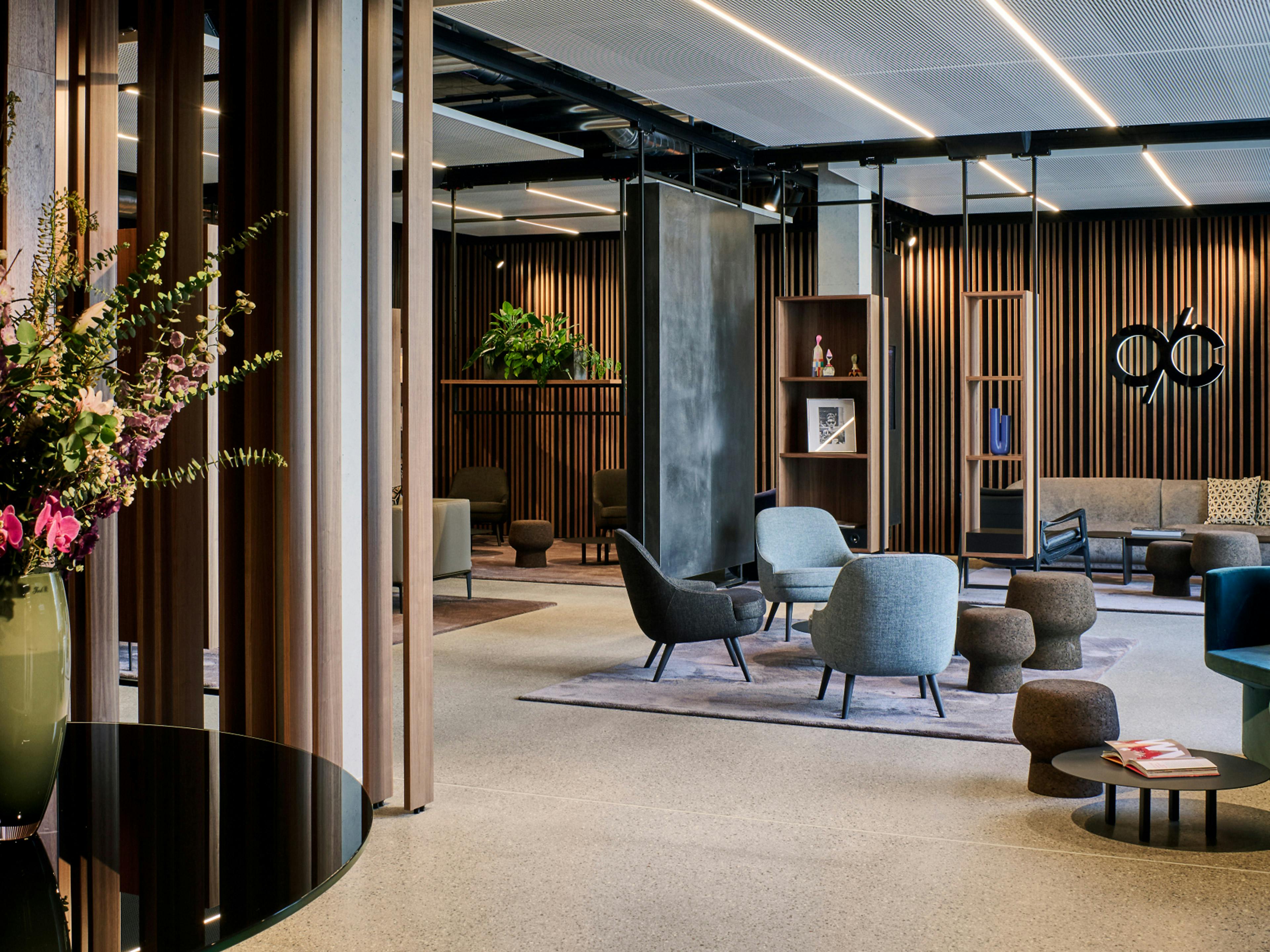 Modern gestaltete Lounge mit gemütlichen Sitzbereichen, stilvollen Sesseln und Hockern auf Teppichen, dekorativen Holzwänden, Pflanzen und einer Blumenvase im Vordergrund auf einem schwarzen Tisch.